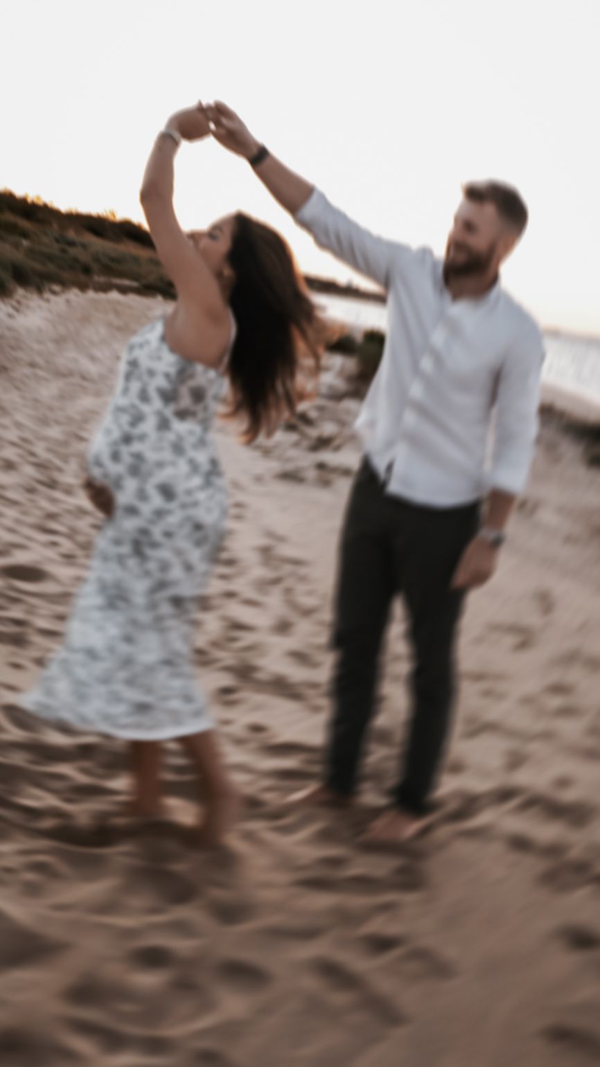 Couple dancing on sandy beach at sunset, man spinning woman in floral dress.