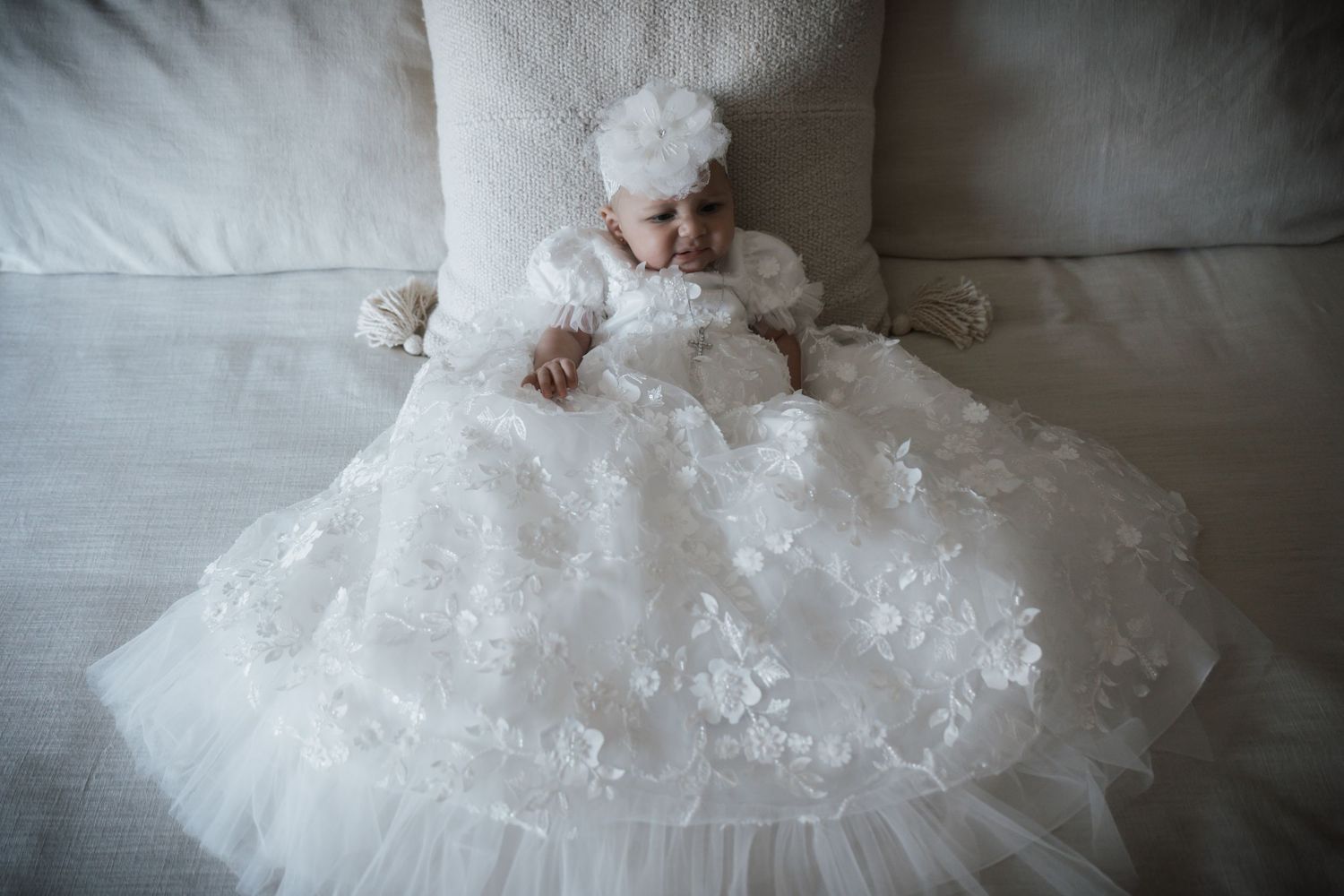 Overhead view of baby in ornate white lace christening gown with floral headband lying on cream sofa.