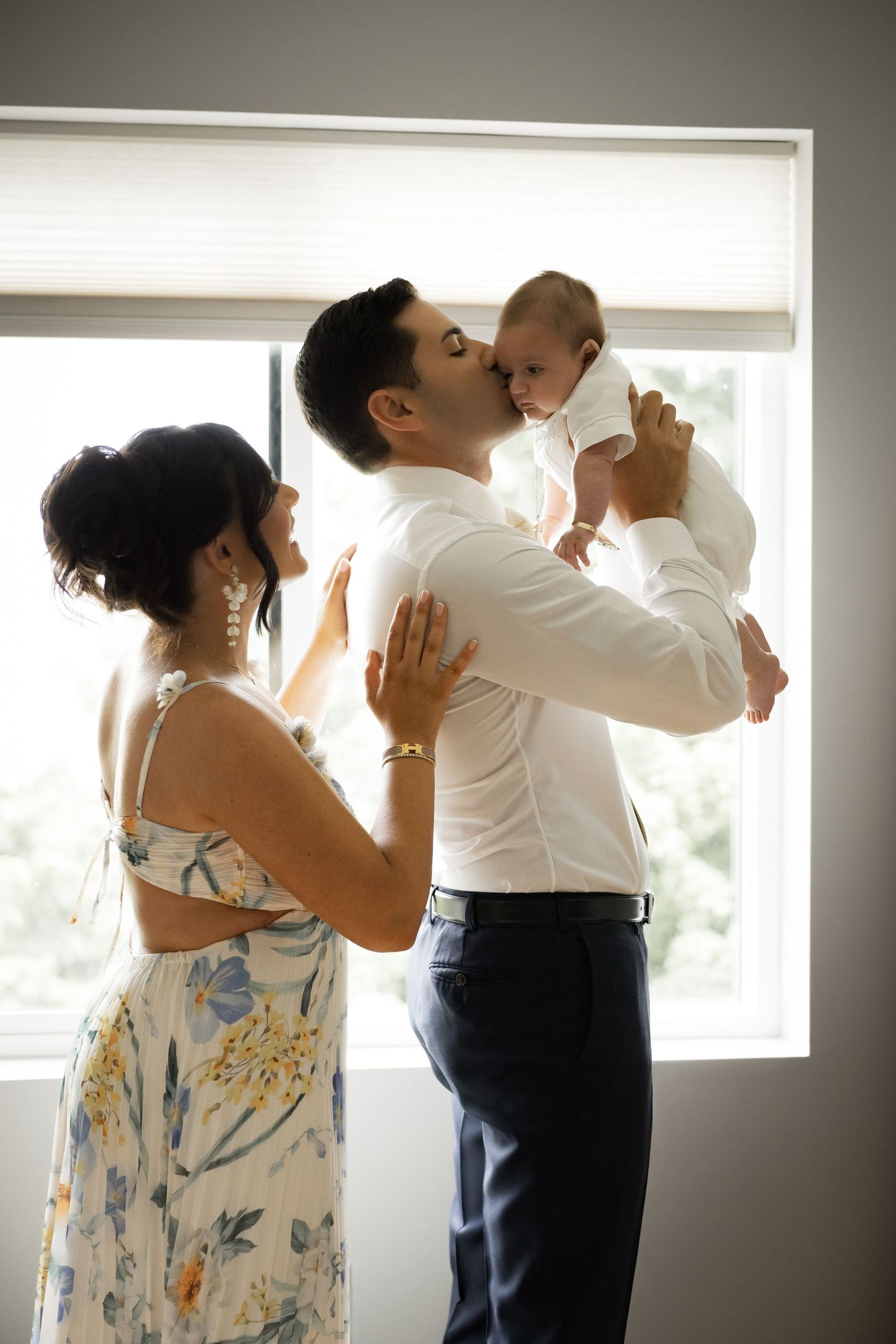 Father kissing newborn baby while mother watches lovingly by window in Staten Island.