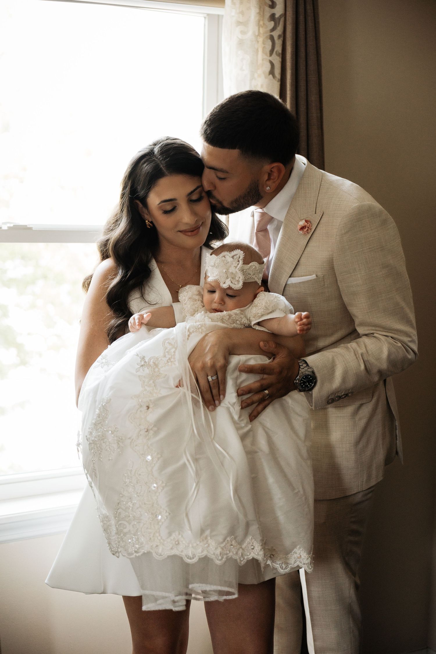 Father in tan suit kissing baby while mother smiles during christening celebration by window.