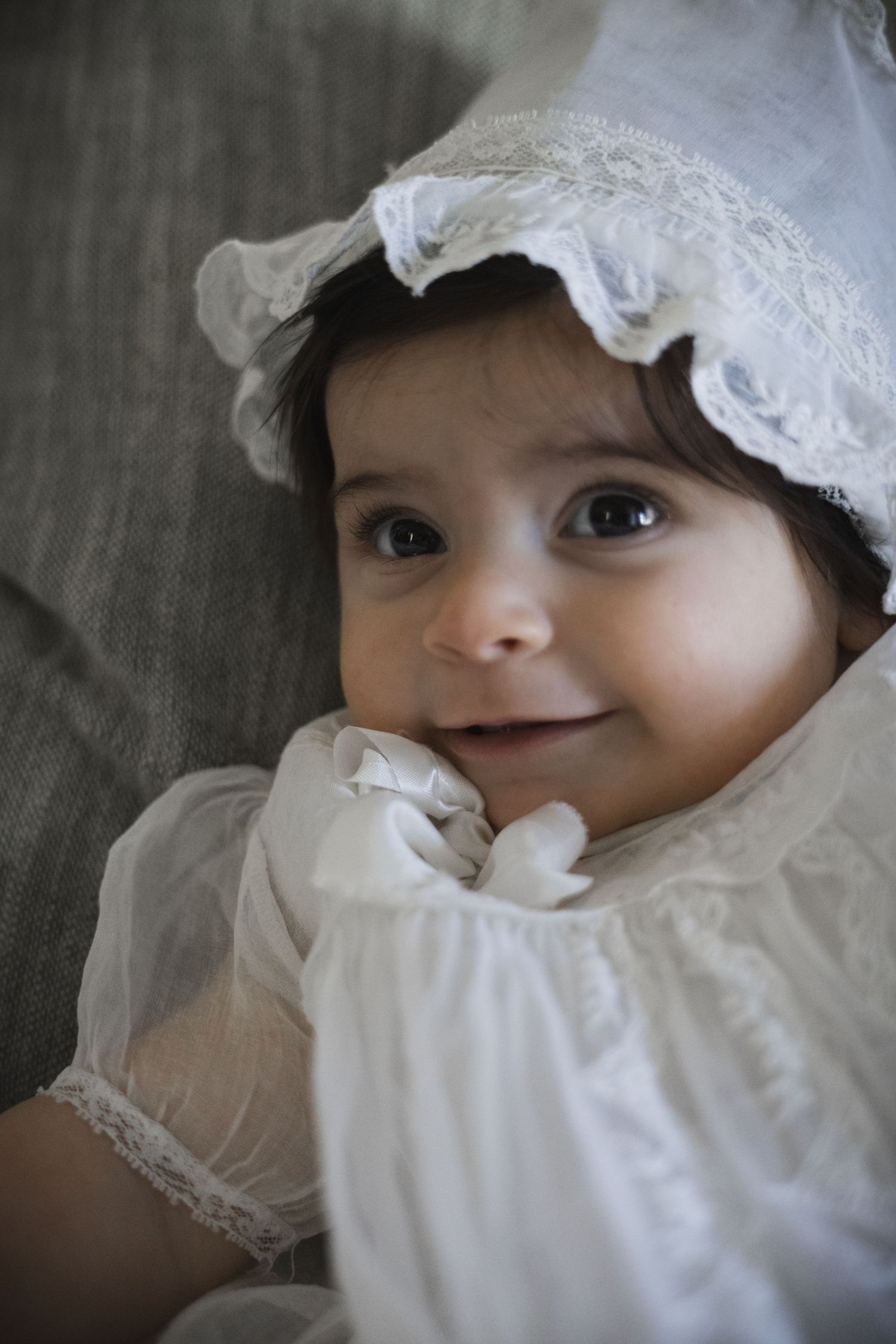 Adorable baby in white lace christening gown and bonnet smiling with bright curious eyes.