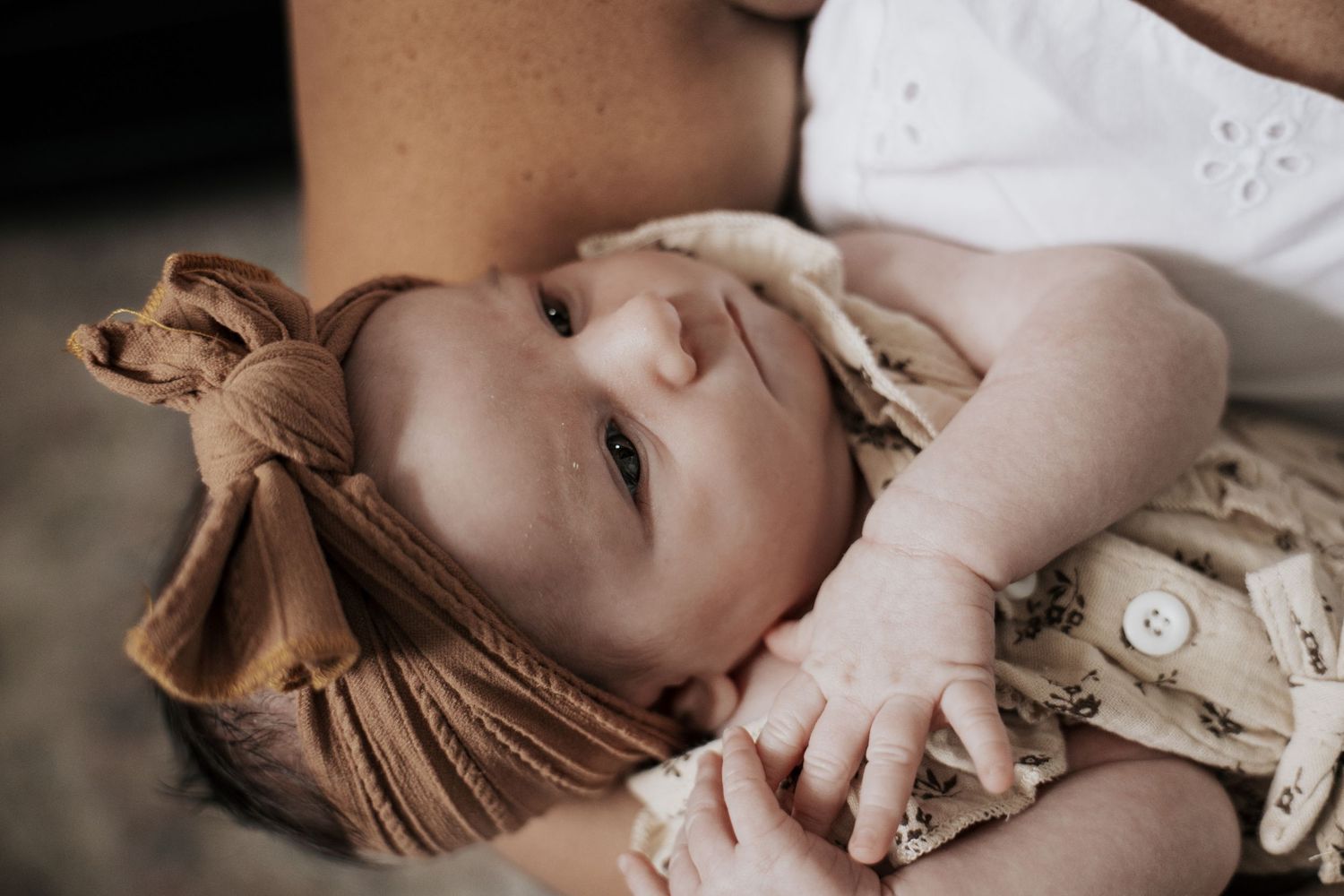 Close-up of newborn baby wearing brown bow headband cradled in mothers arms in soft natural light.