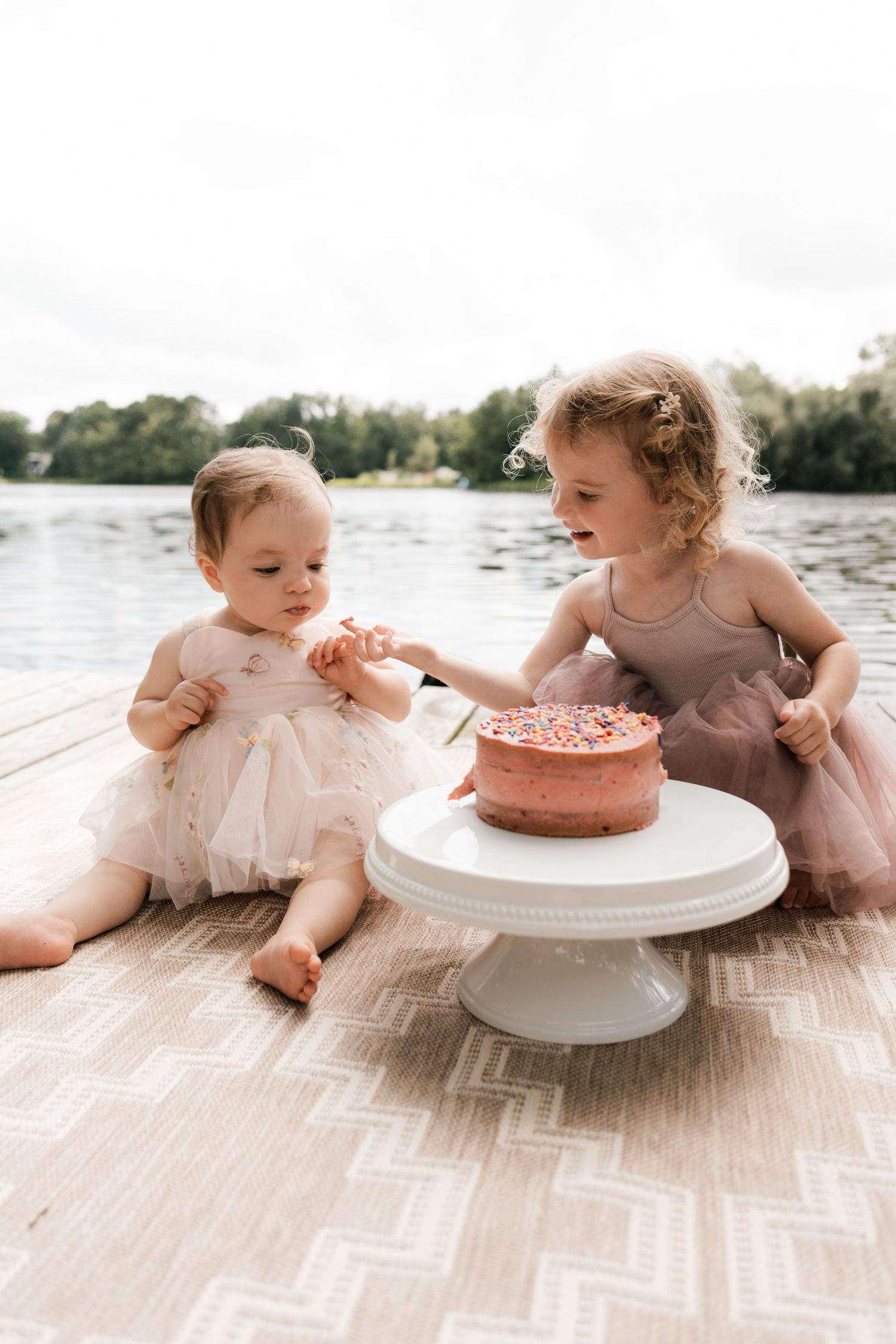 Two little sisters in pink dresses sit on dock with birthday cake by lake.