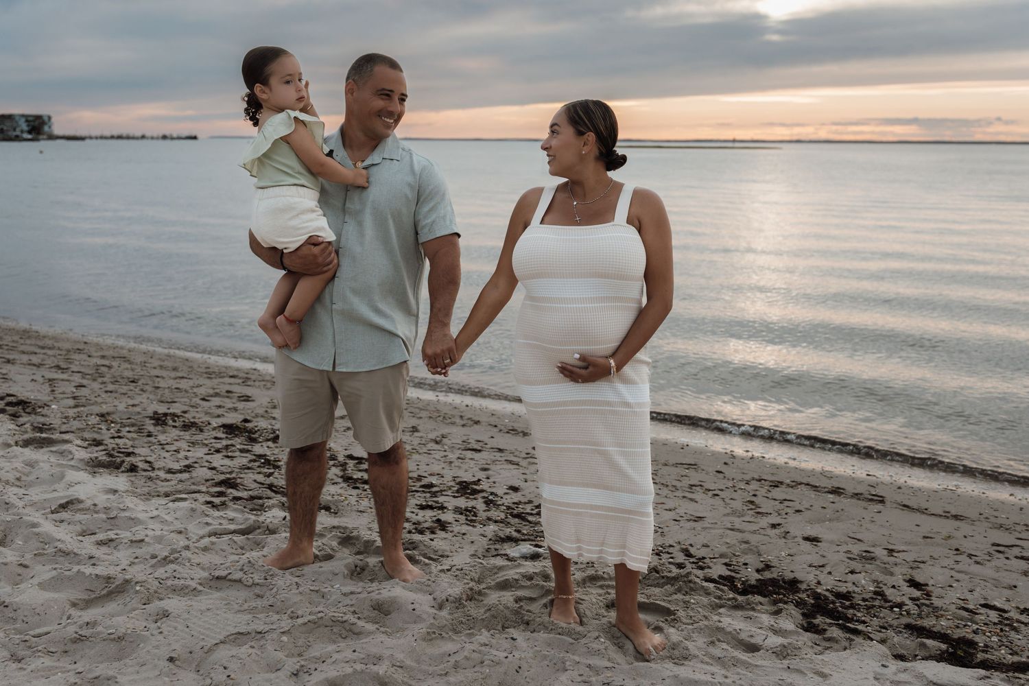 Pregnant woman in white dress holds hands with man carrying toddler on sandy beach at sunset.