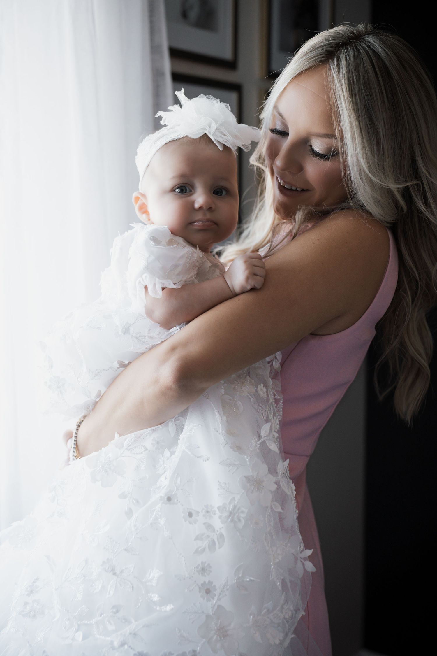 Smiling blonde woman in pink dress holding baby dressed in elaborate white lace christening gown by window.