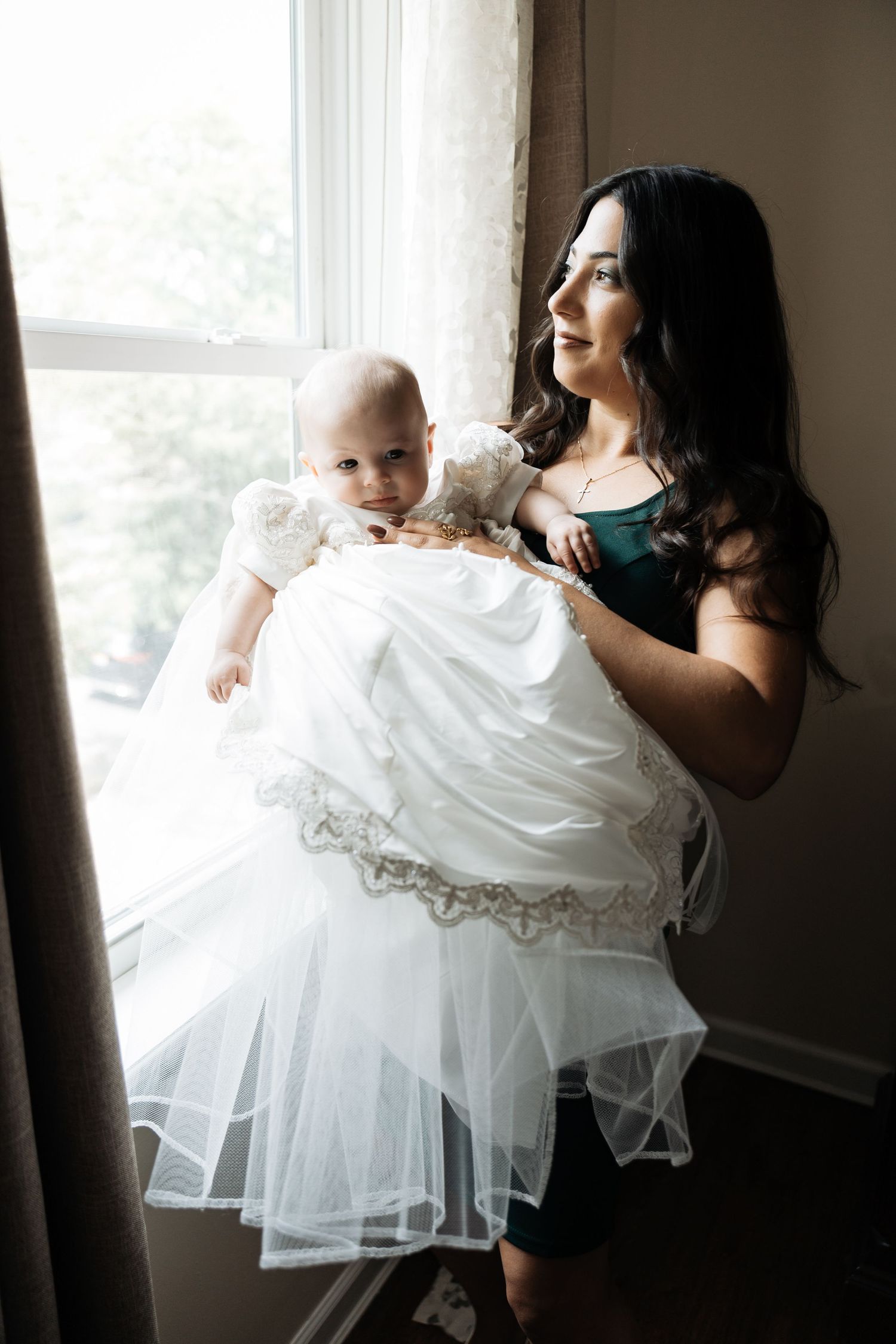 Woman in green dress holding baby in white christening gown while gazing out window.