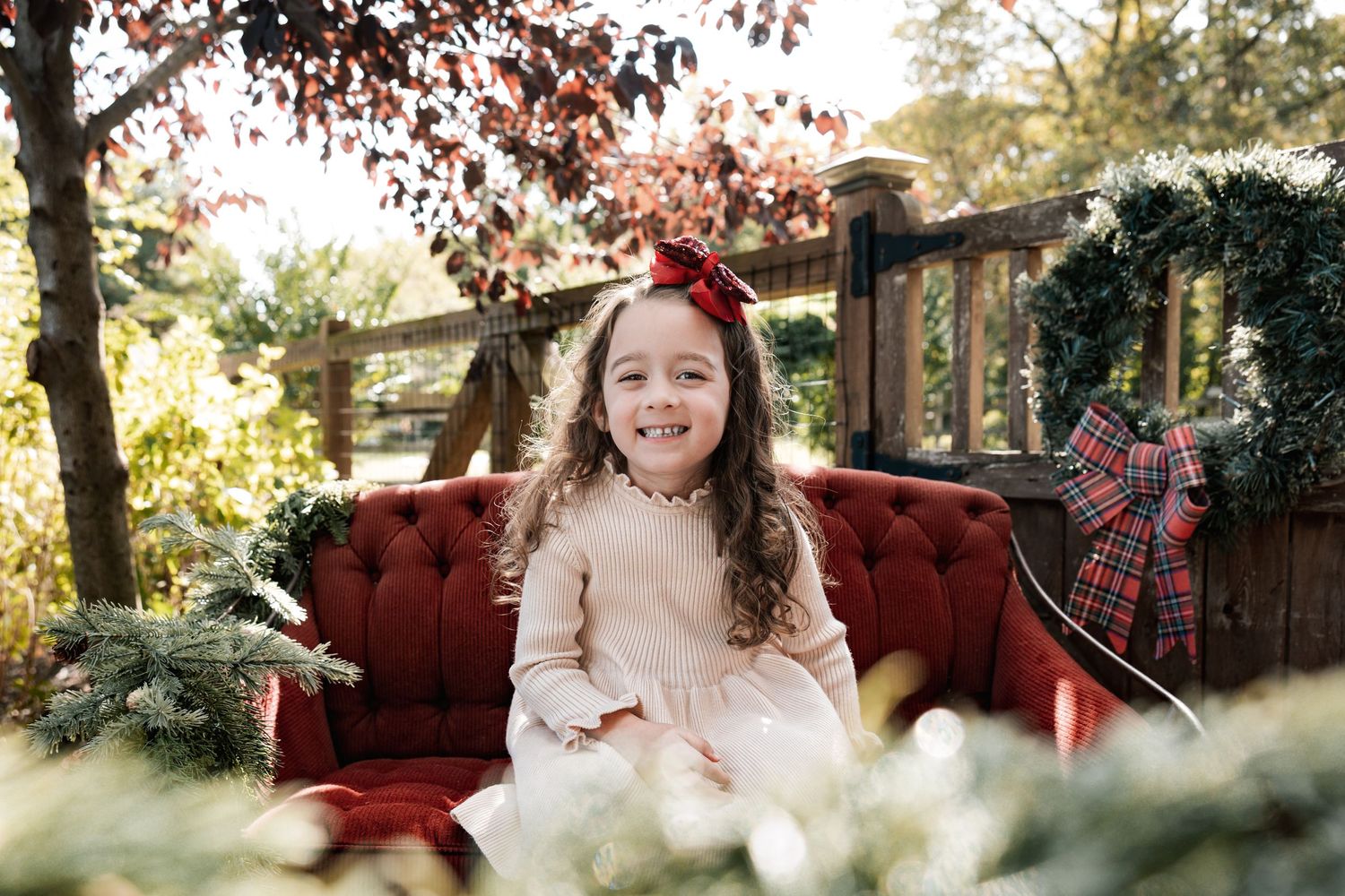 A cheerful young girl in a cream dress sits on a red velvet sofa surrounded by Christmas greenery outdoors.