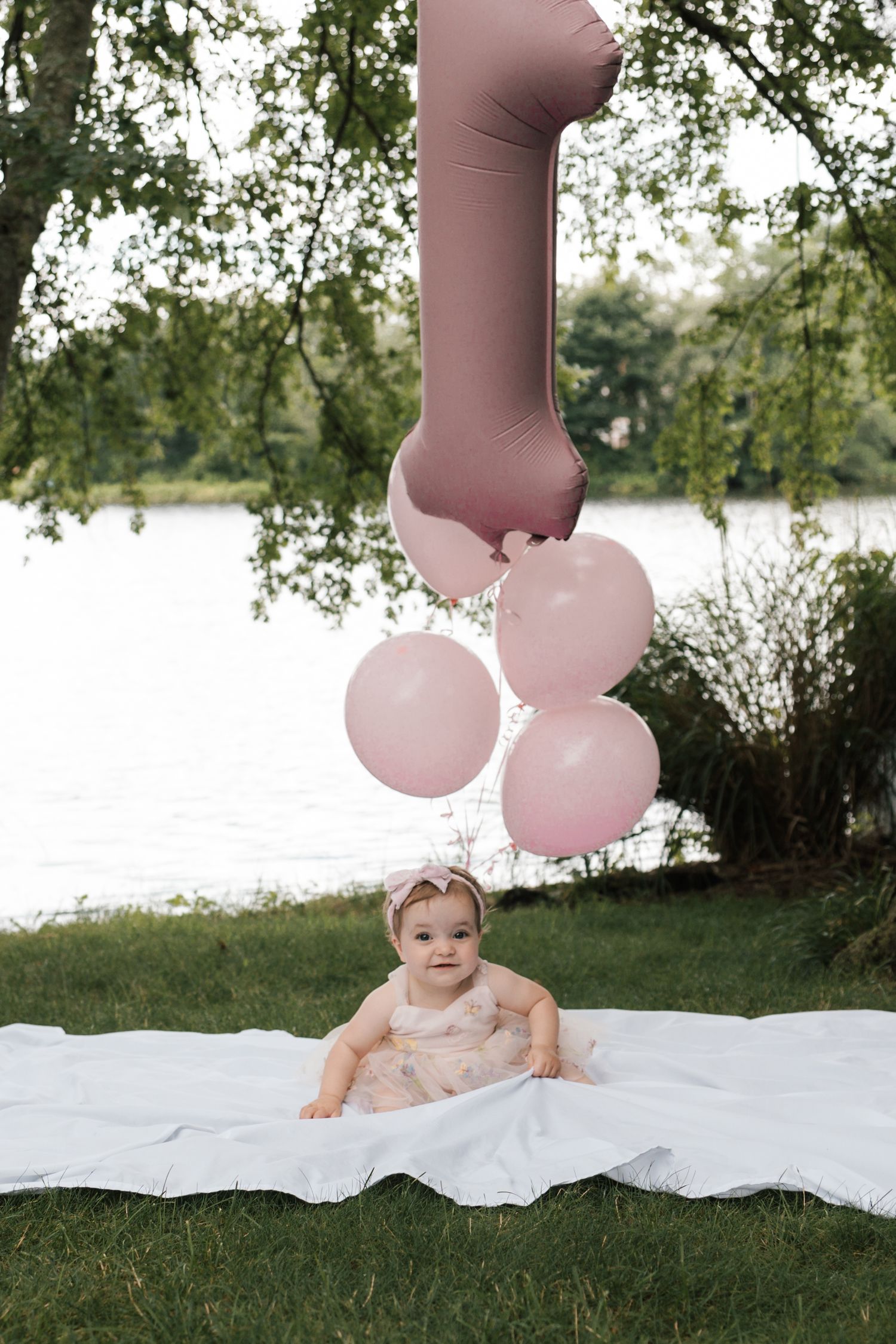 Baby girl in floral dress sitting on white blanket outdoors with pink number one balloon bouquet.