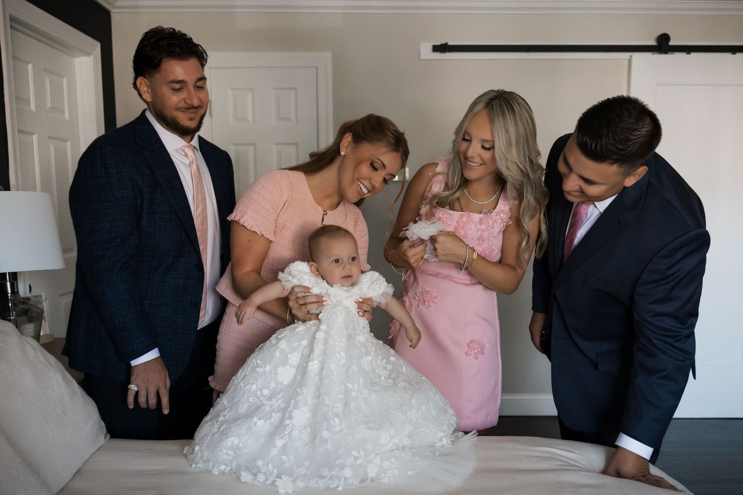Group of elegantly dressed family members gathered around baby in elaborate white lace christening gown indoors.
