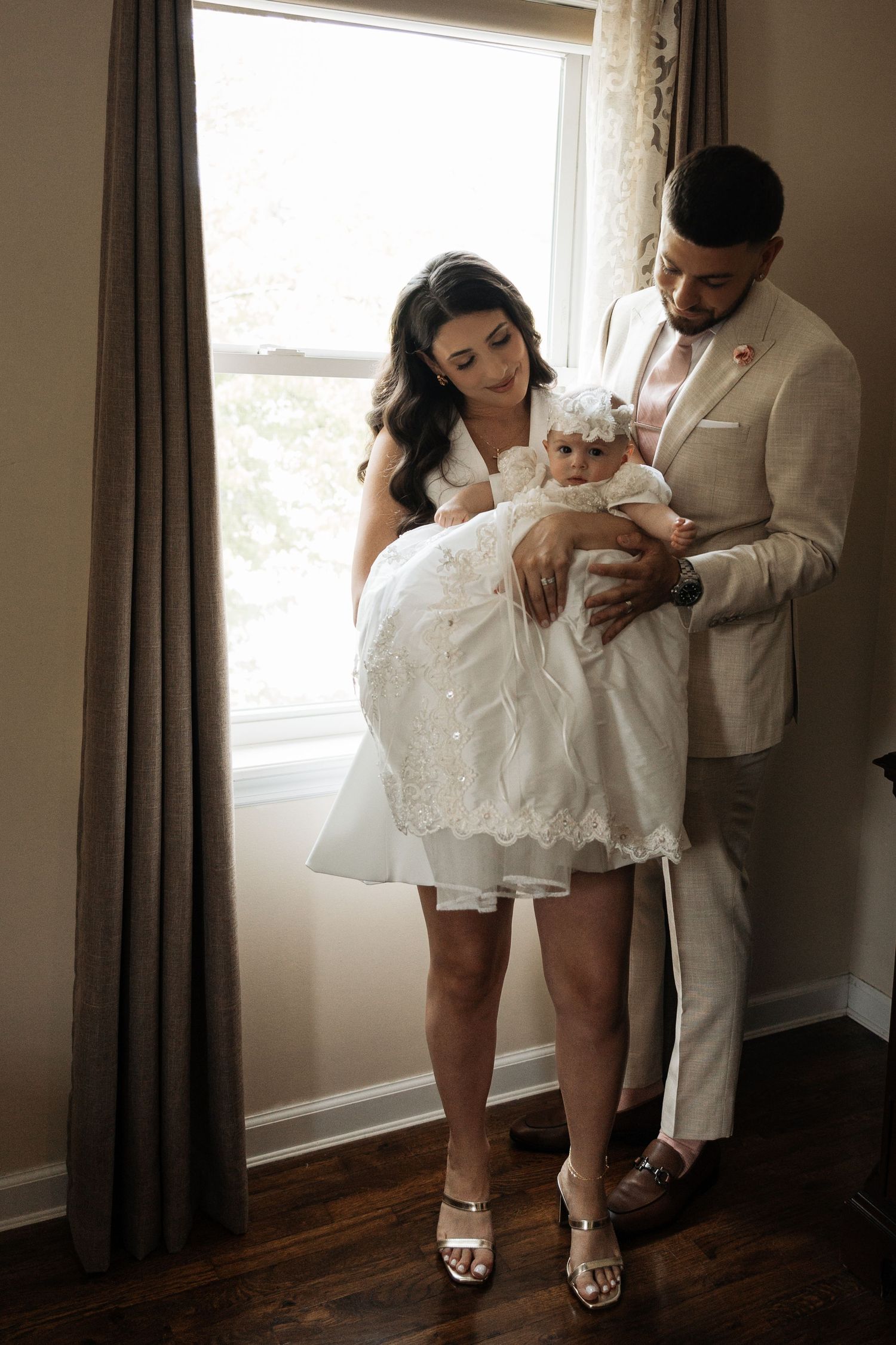 Parents in formal attire lovingly holding baby in white christening gown by a window.