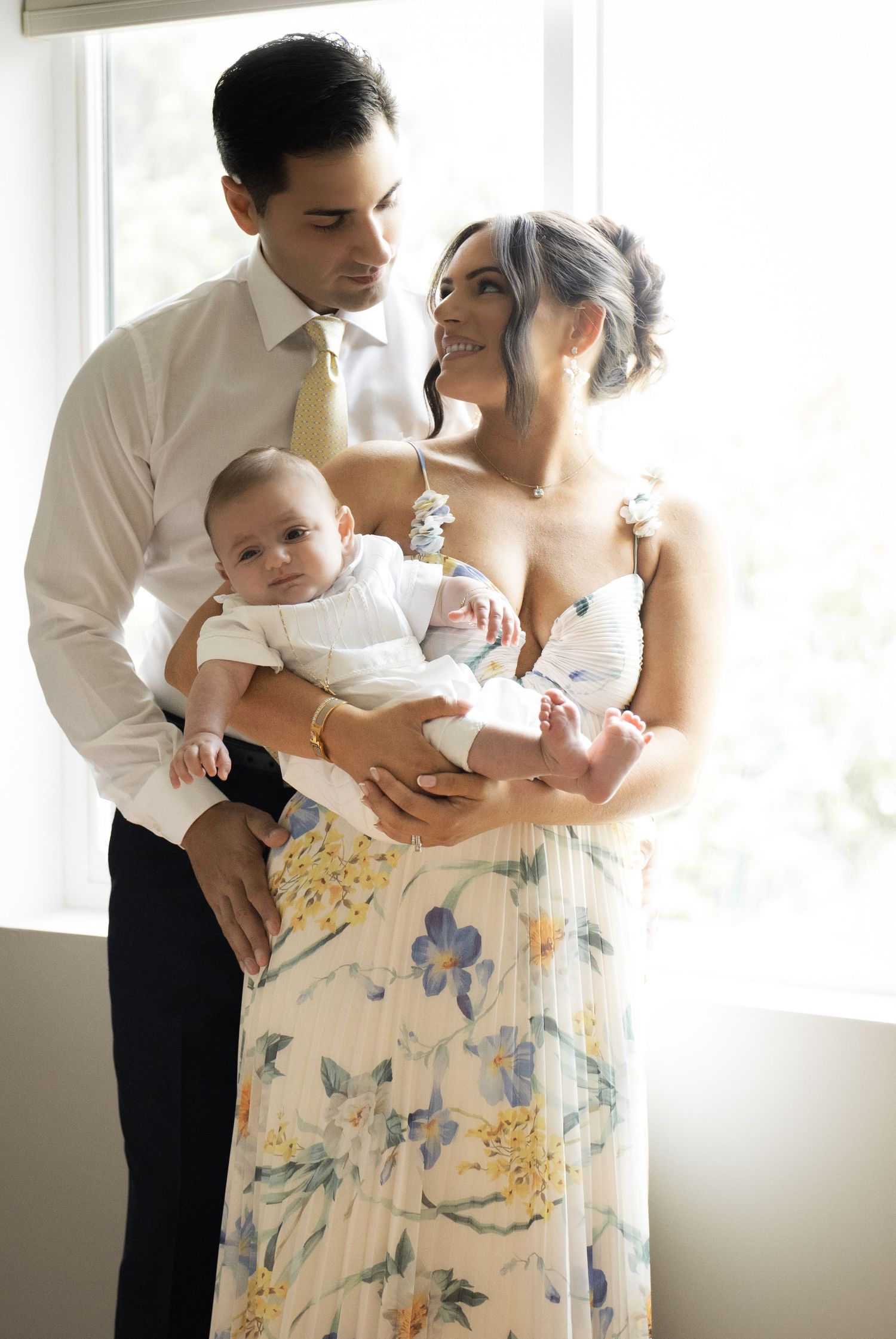 Young couple in formal attire holding newborn twins by a bright window In Staten Island.