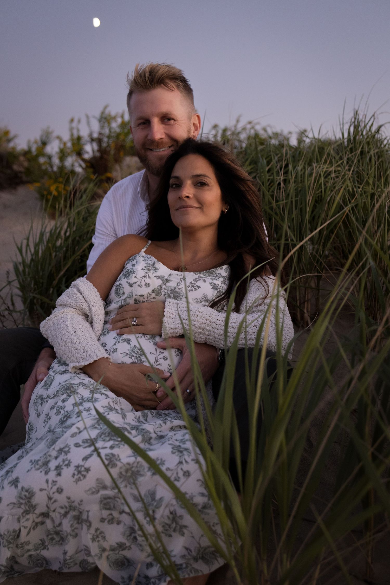 Couple sitting together in beach grass at dusk with moon visible, woman in floral maternity dress.