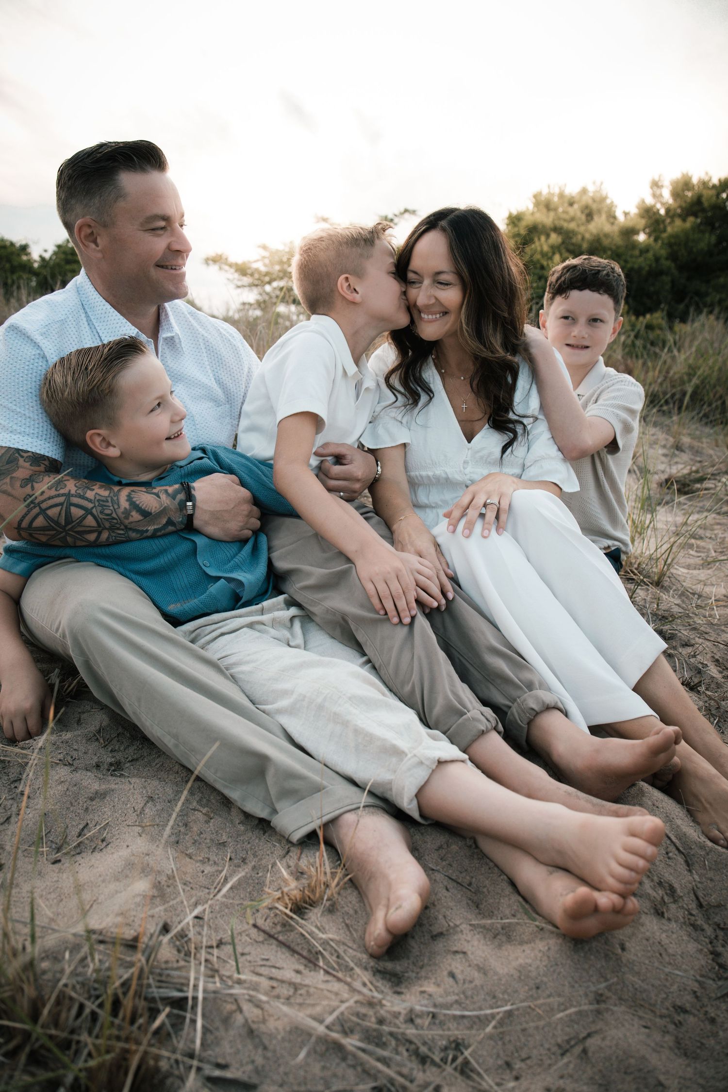 A happy family of five sits together on sandy dunes at sunset, laughing and cuddling.