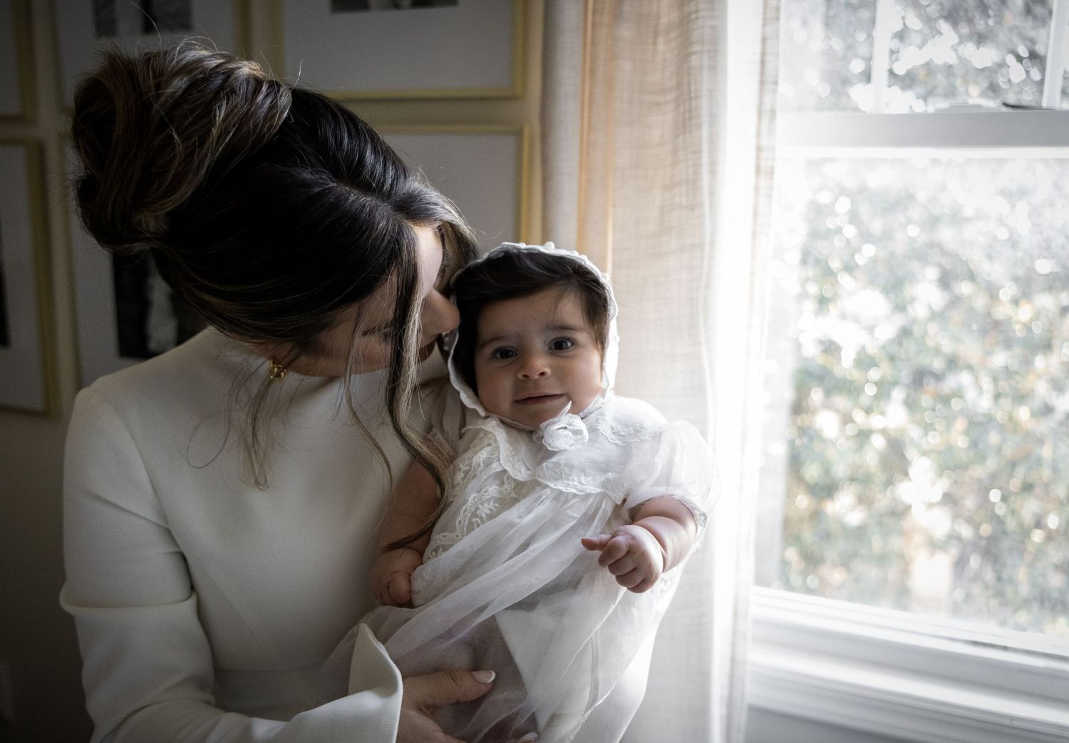 Mother tenderly kissing her wide-eyed baby dressed in a white christening gown by a bright window.