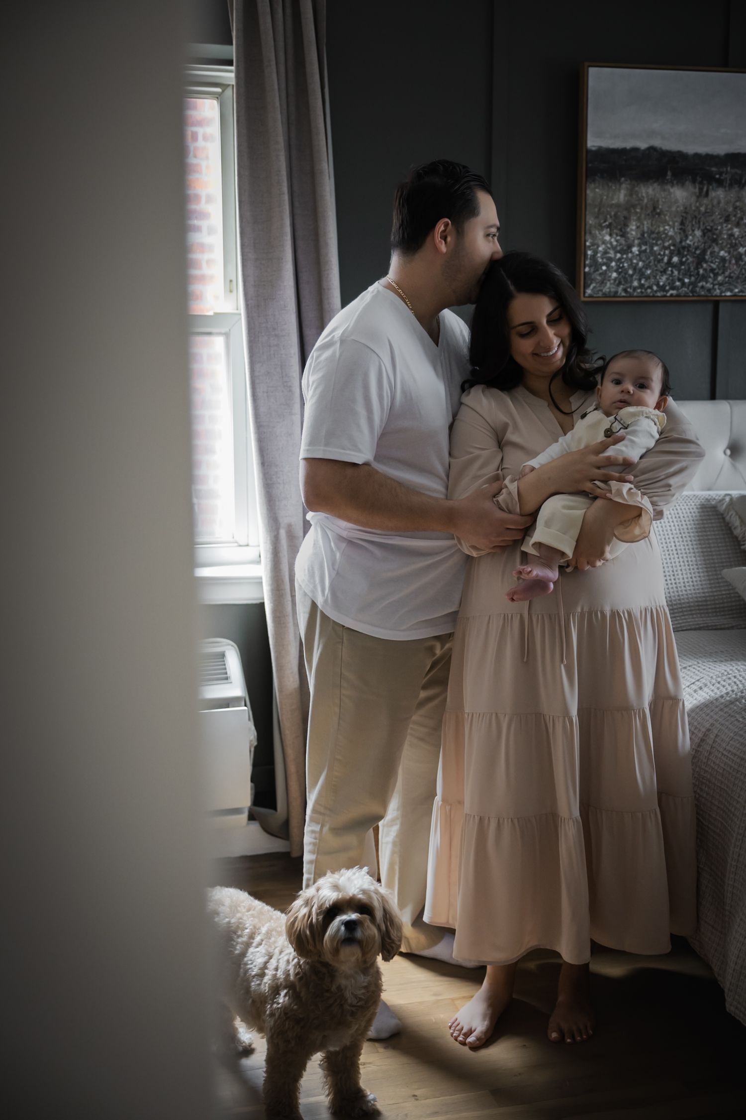 A young family with a newborn baby and fluffy dog pose together in a cozy bedroom near a window.