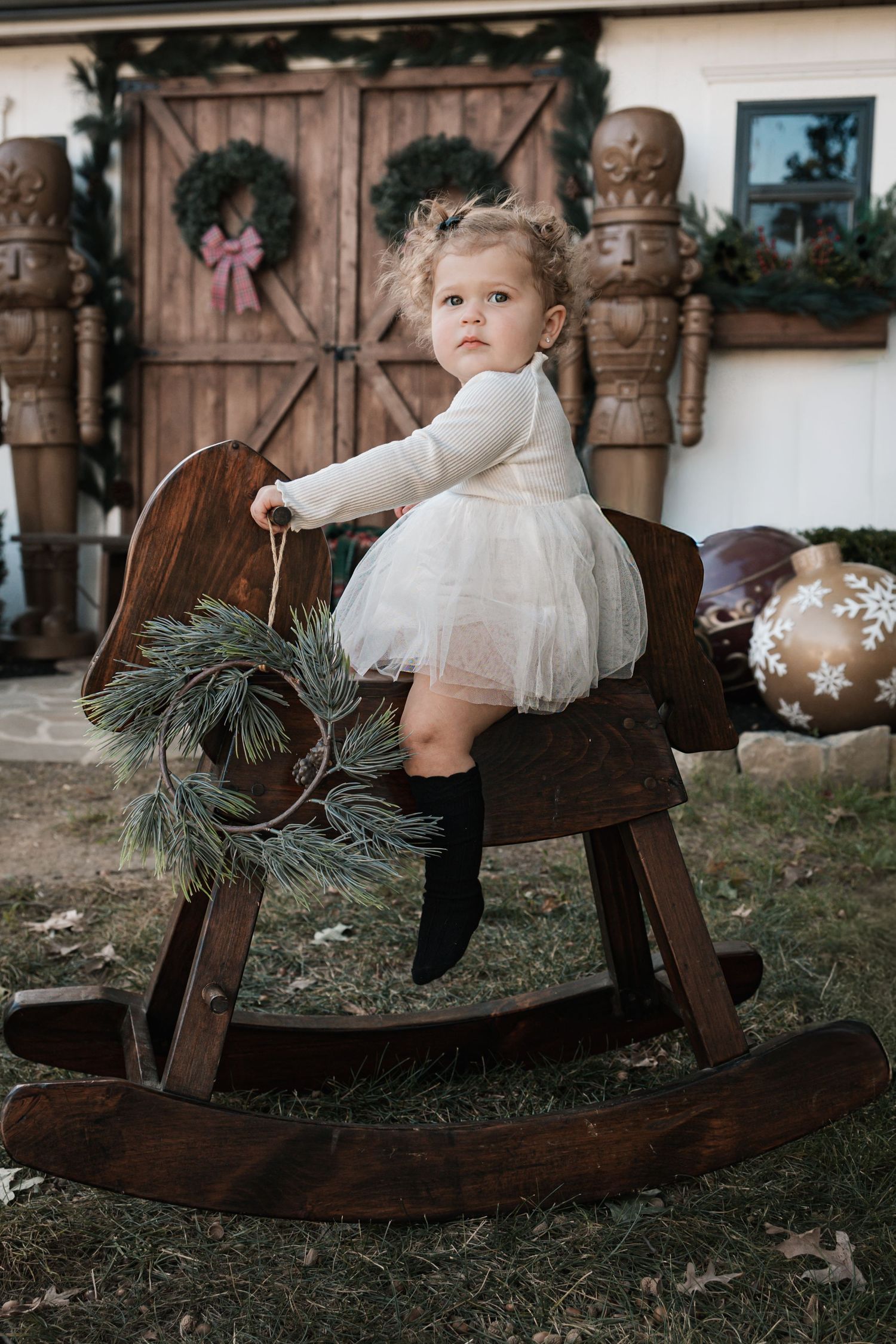 Toddler girl in white dress and black boots riding a vintage wooden rocking horse outdoors at Christmas.