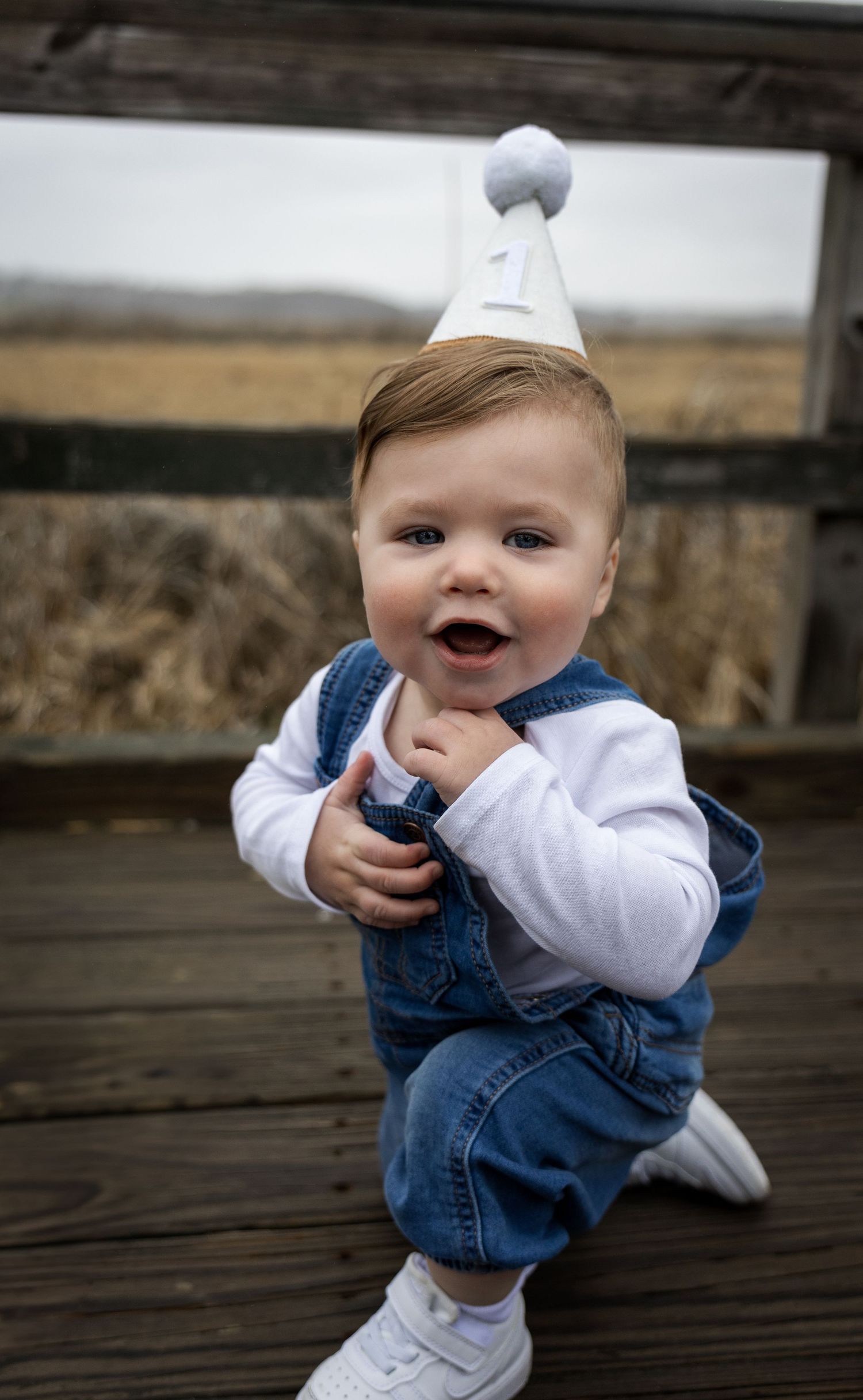 Happy baby boy in denim overalls and white birthday party hat kneeling on wooden deck.