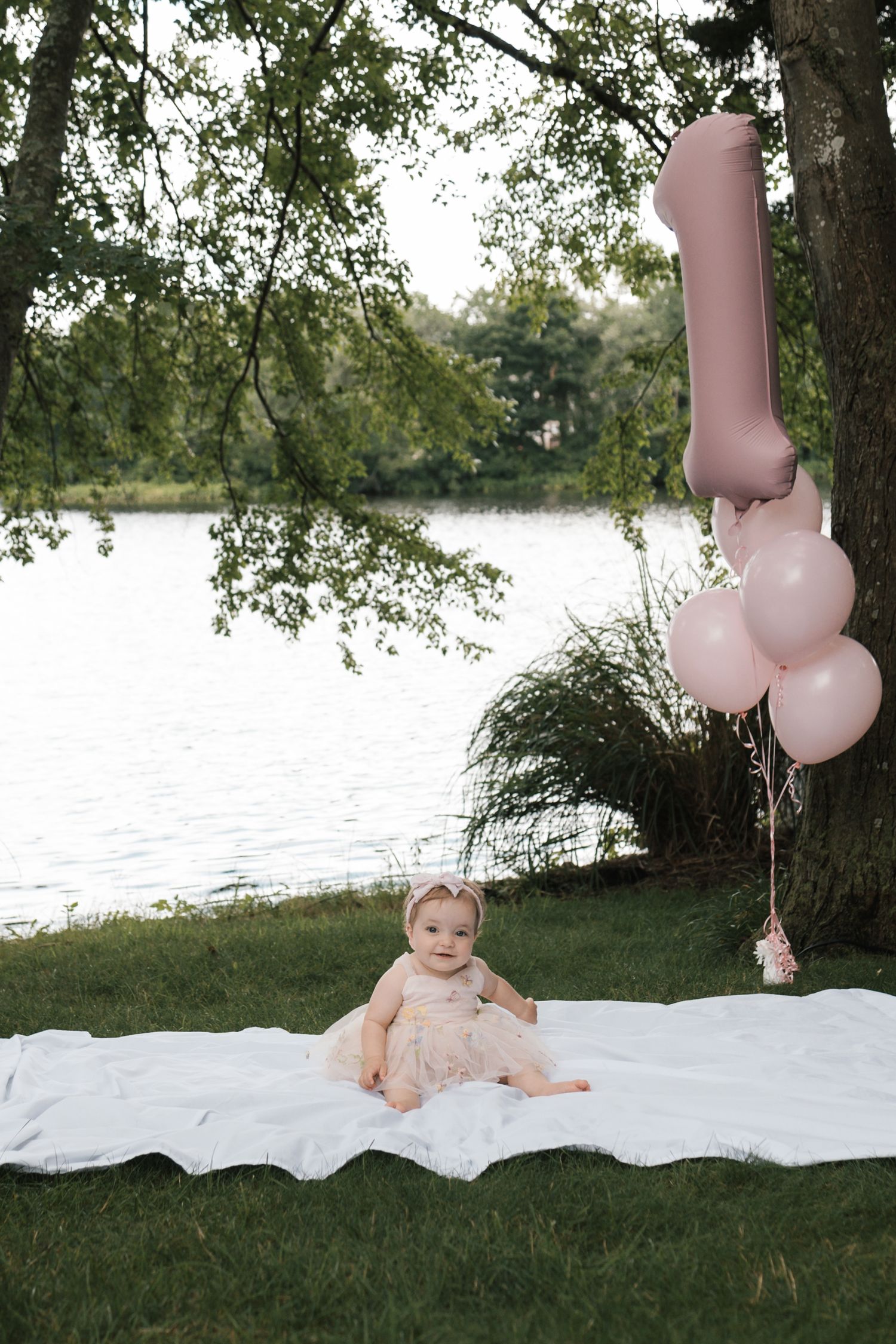 Baby girl in pink dress celebrating first birthday with balloons by lake in Staten Island.