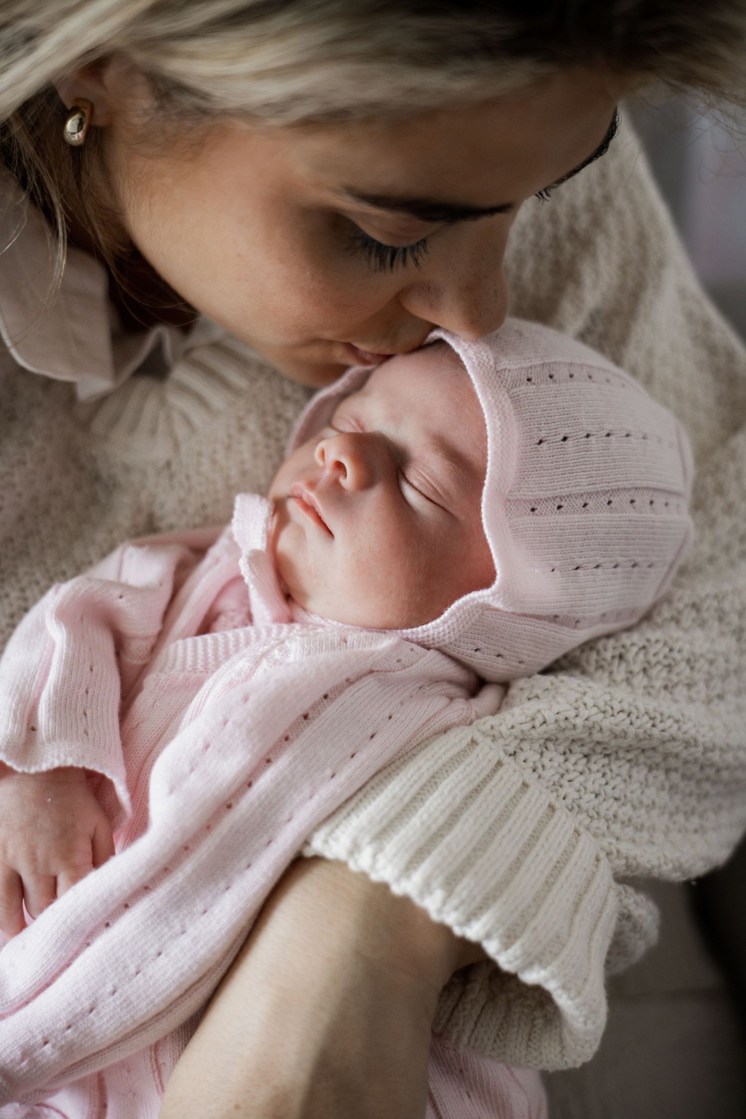 Mother gently kisses sleeping newborn dressed in pink knit outfit and hat.