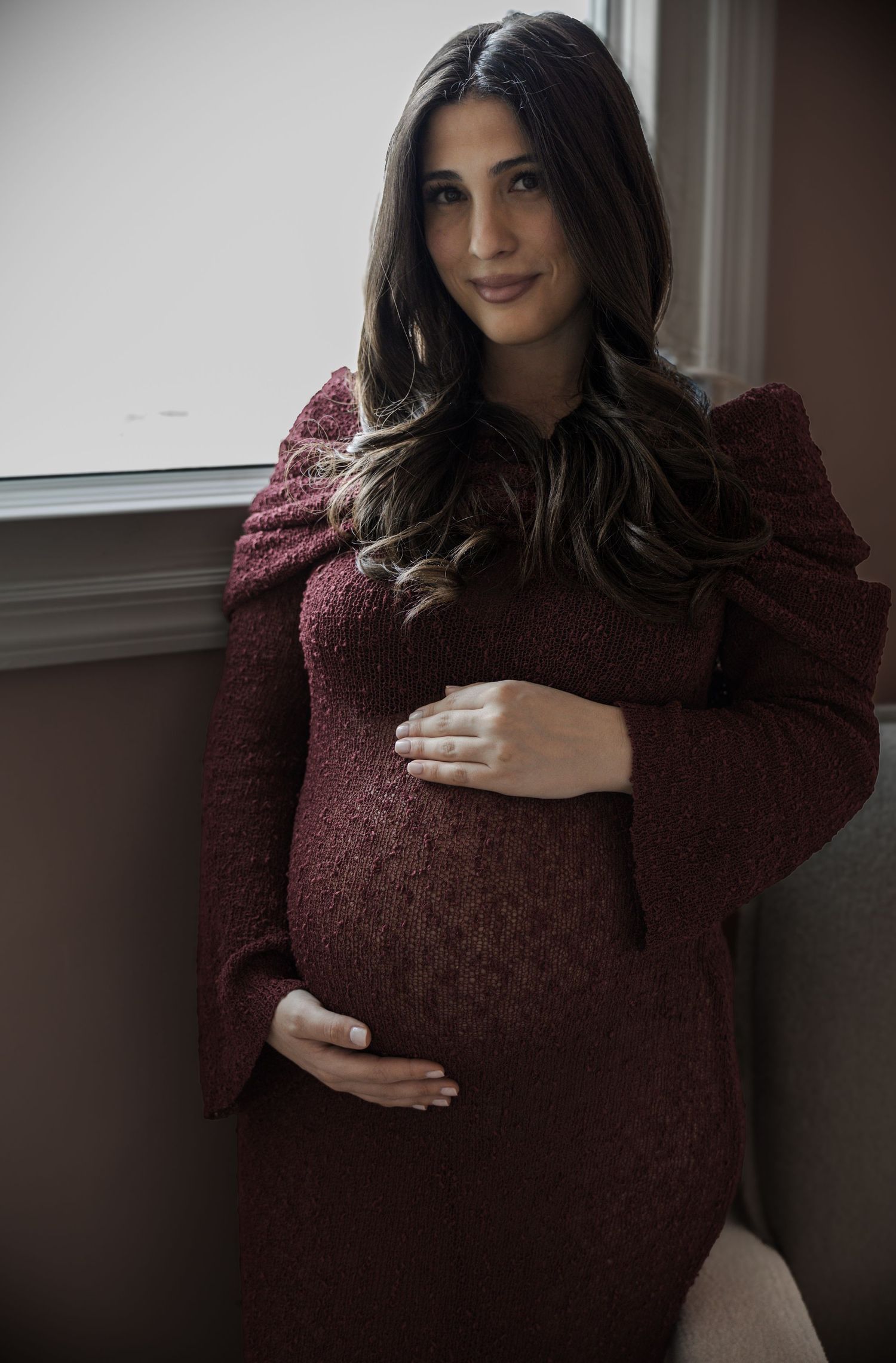 Smiling pregnant woman in burgundy knit dress holding baby bump near window for maternity portrait.