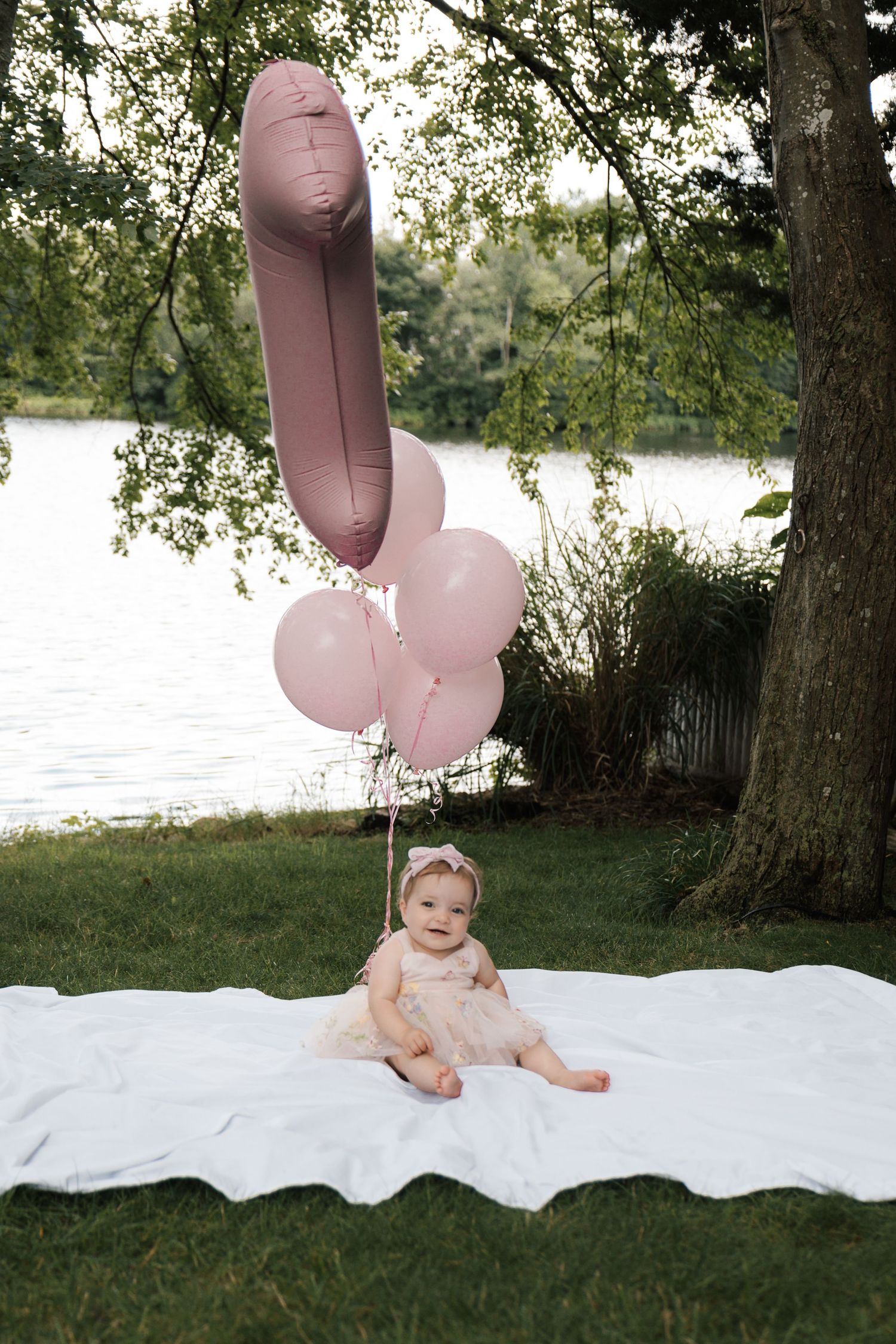 An adorable baby girl in a pink dress sits on a white blanket outdoors with pink balloons by a lake in New Jersey.