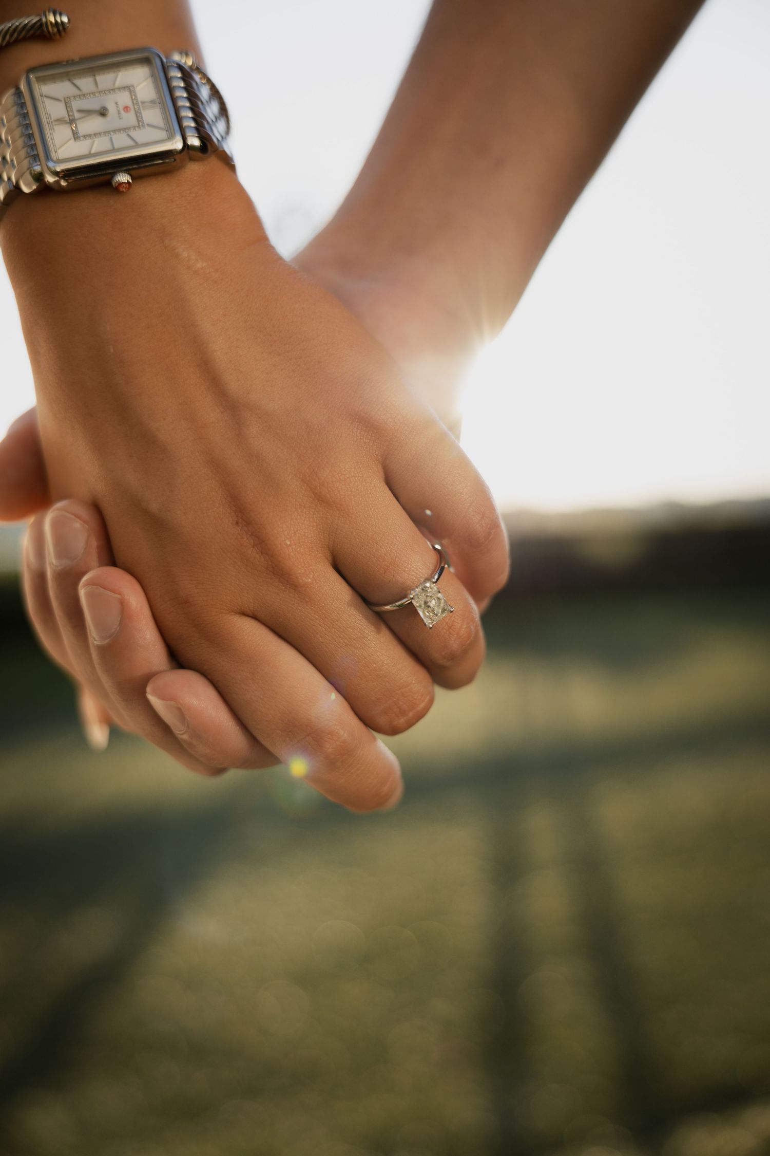 Close-up of couple holding hands in sunlight, showcasing a sparkling square diamond engagement ring.