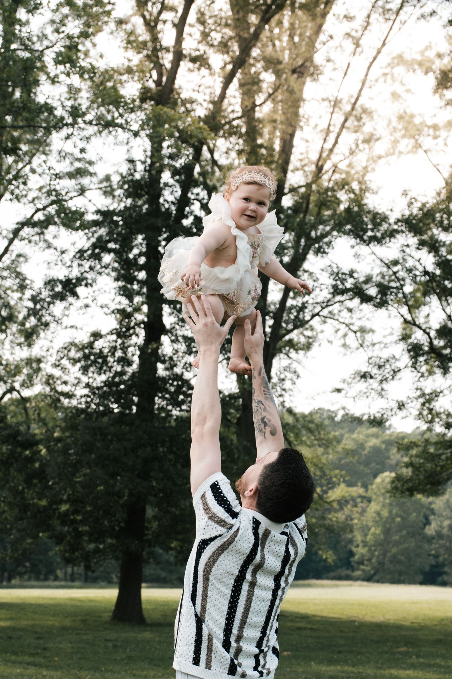 Father tosses laughing baby girl in white dress into air in sunny park.