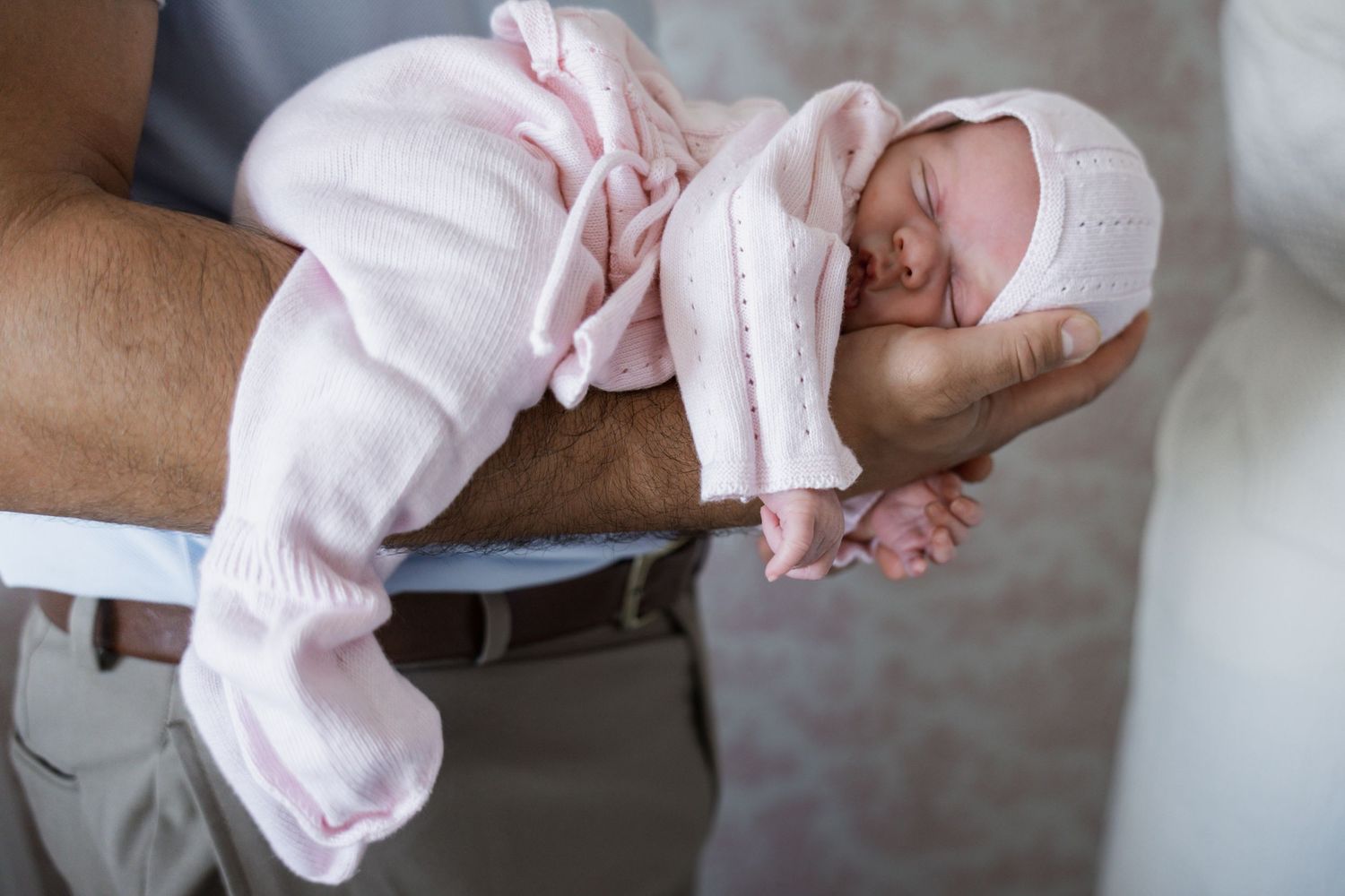 Newborn baby in pink knit outfit sleeps peacefully in fathers arms.