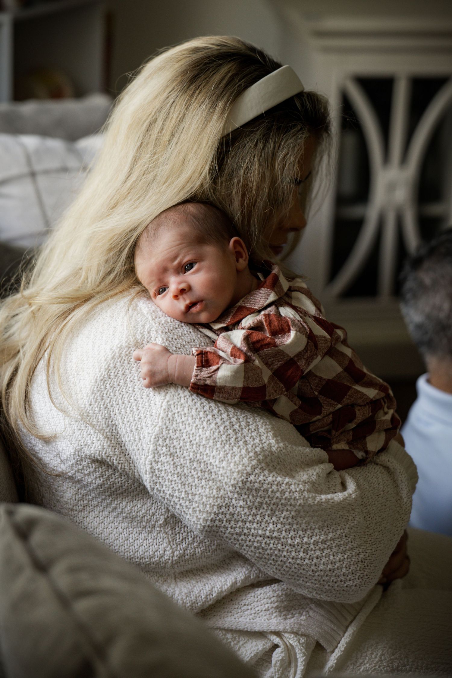 Blonde mother in white sweater gently holds sleeping newborn baby against her shoulder.