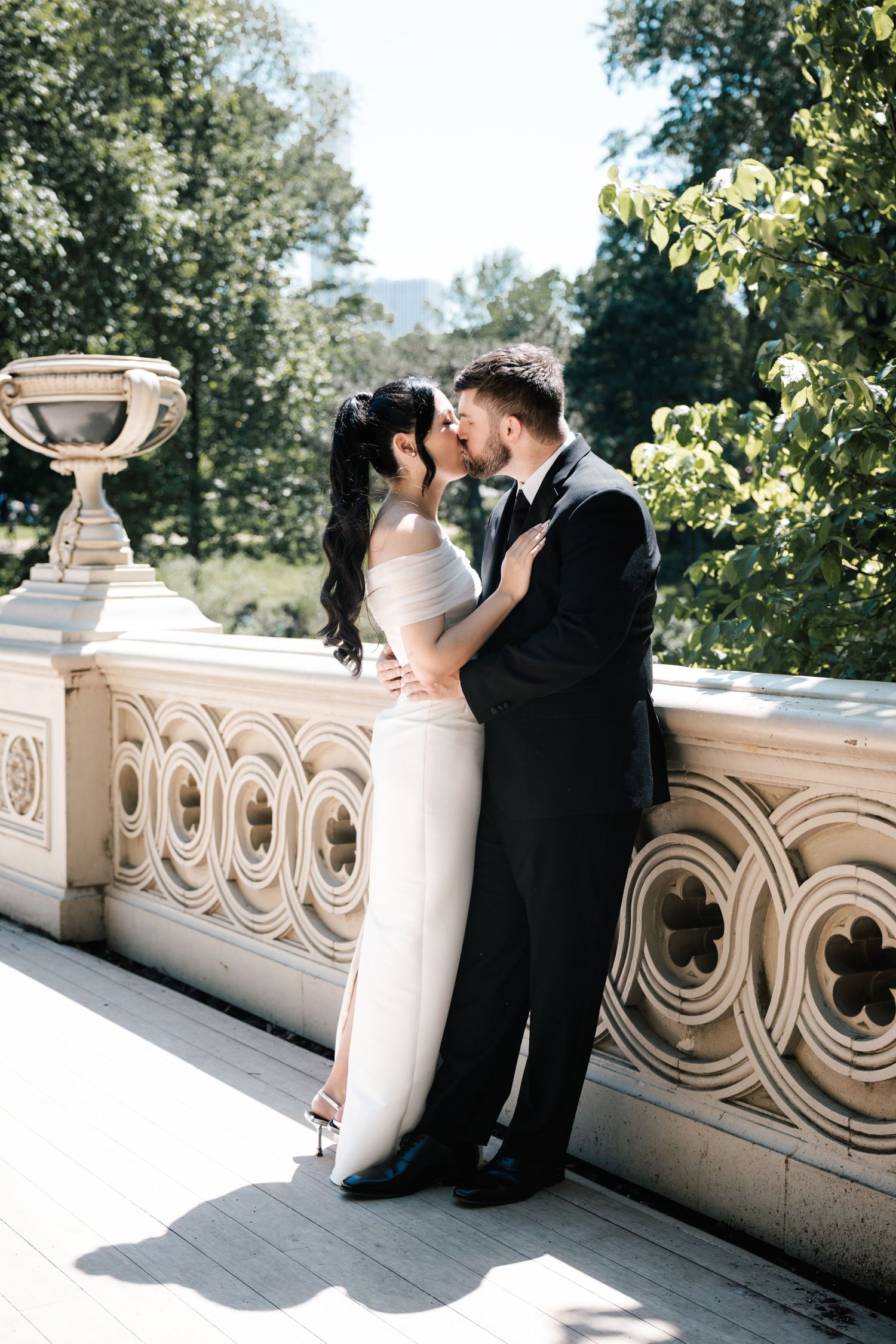 Bride and groom share a tender kiss on ornate stone terrace surrounded by lush green trees in sunlight in New Jersey.