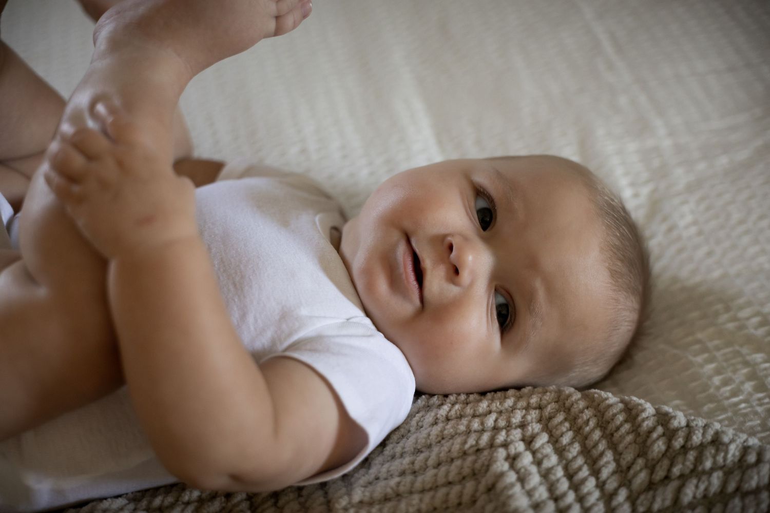 Happy baby in white onesie lying on back smiling while playing with feet on beige blanket.