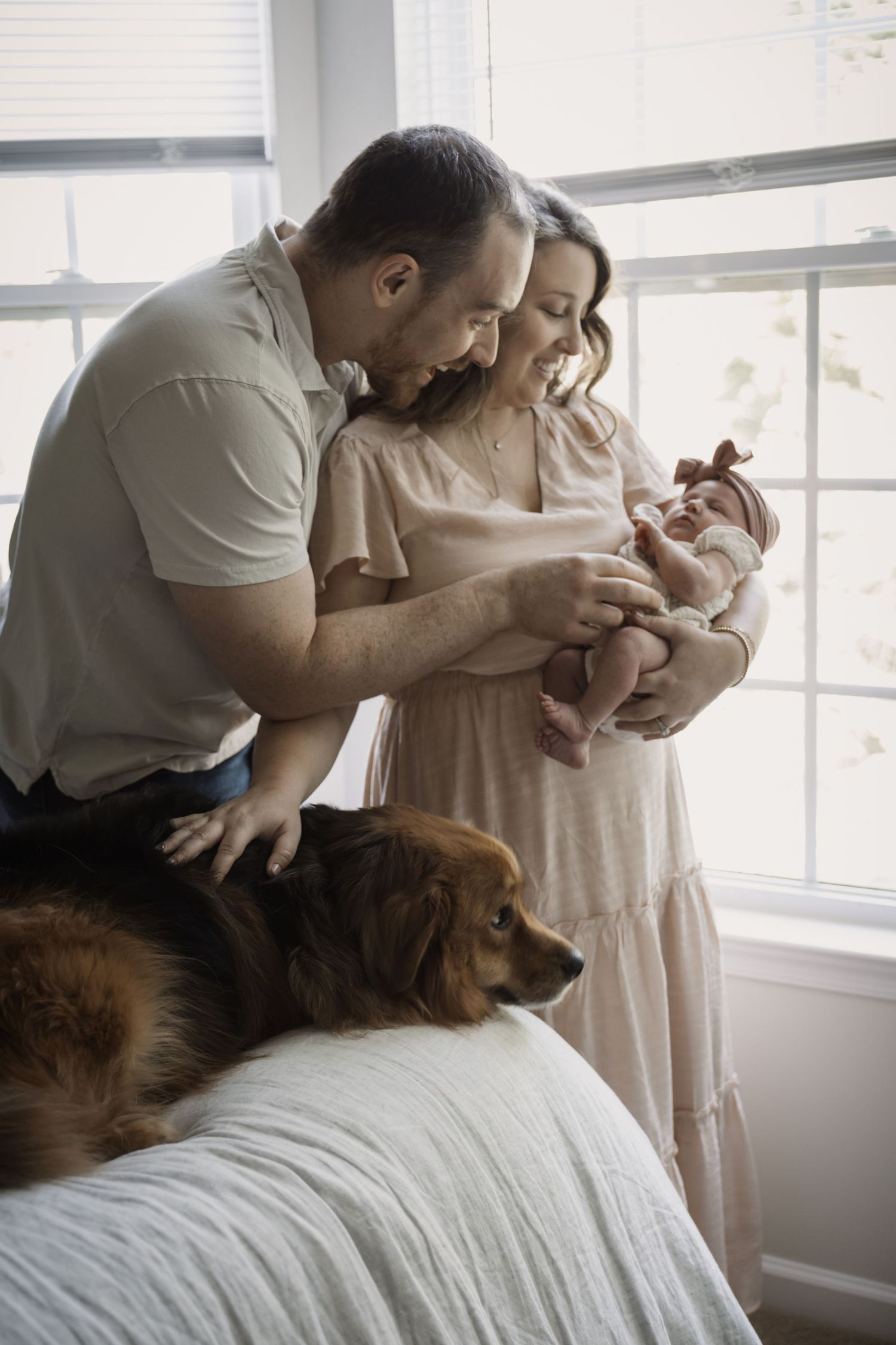 Happy parents smile at their newborn baby girl while their golden retriever watches nearby.