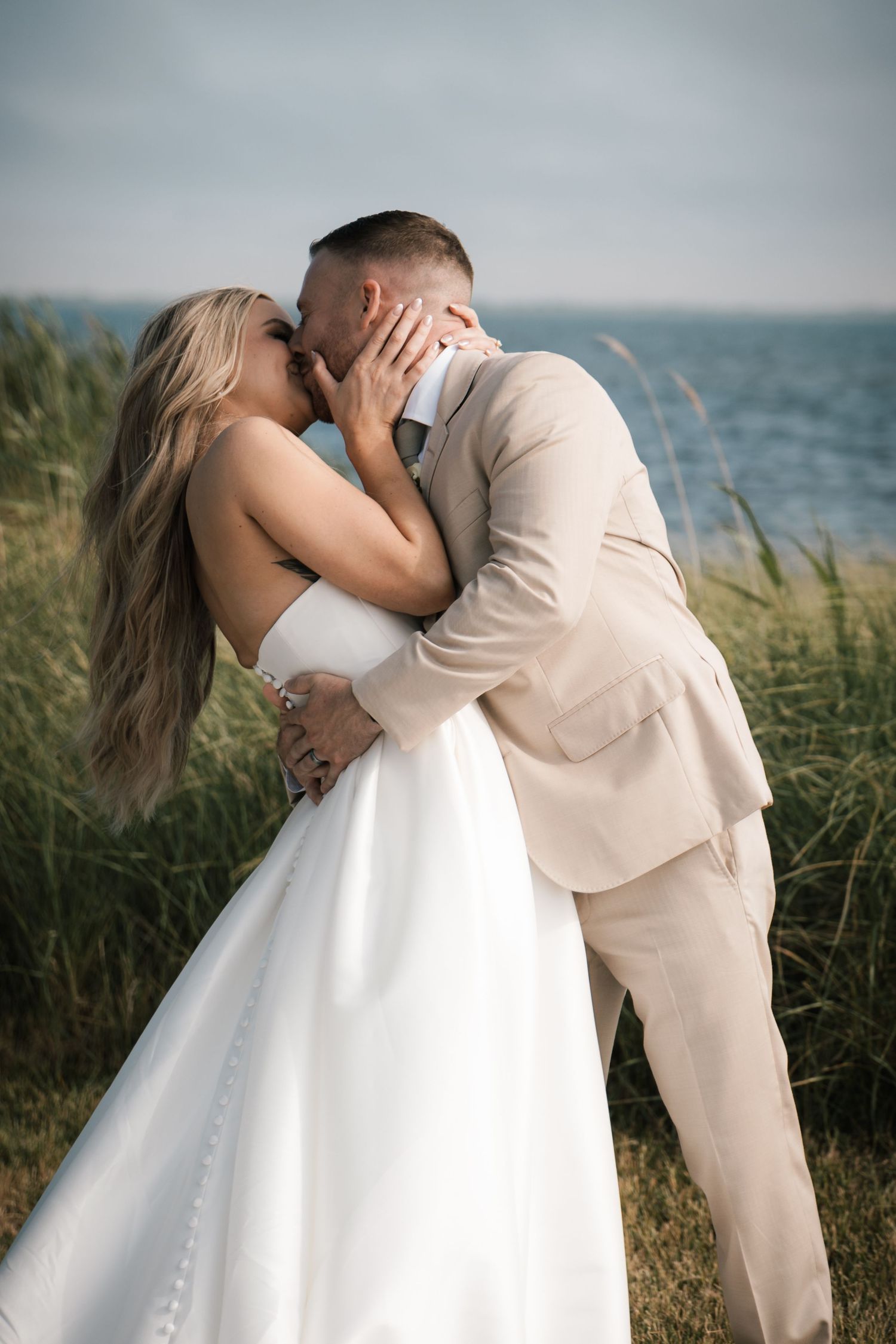 Bride and groom share a romantic kiss near coastal tall grass with ocean water visible in the background in New Jersey.