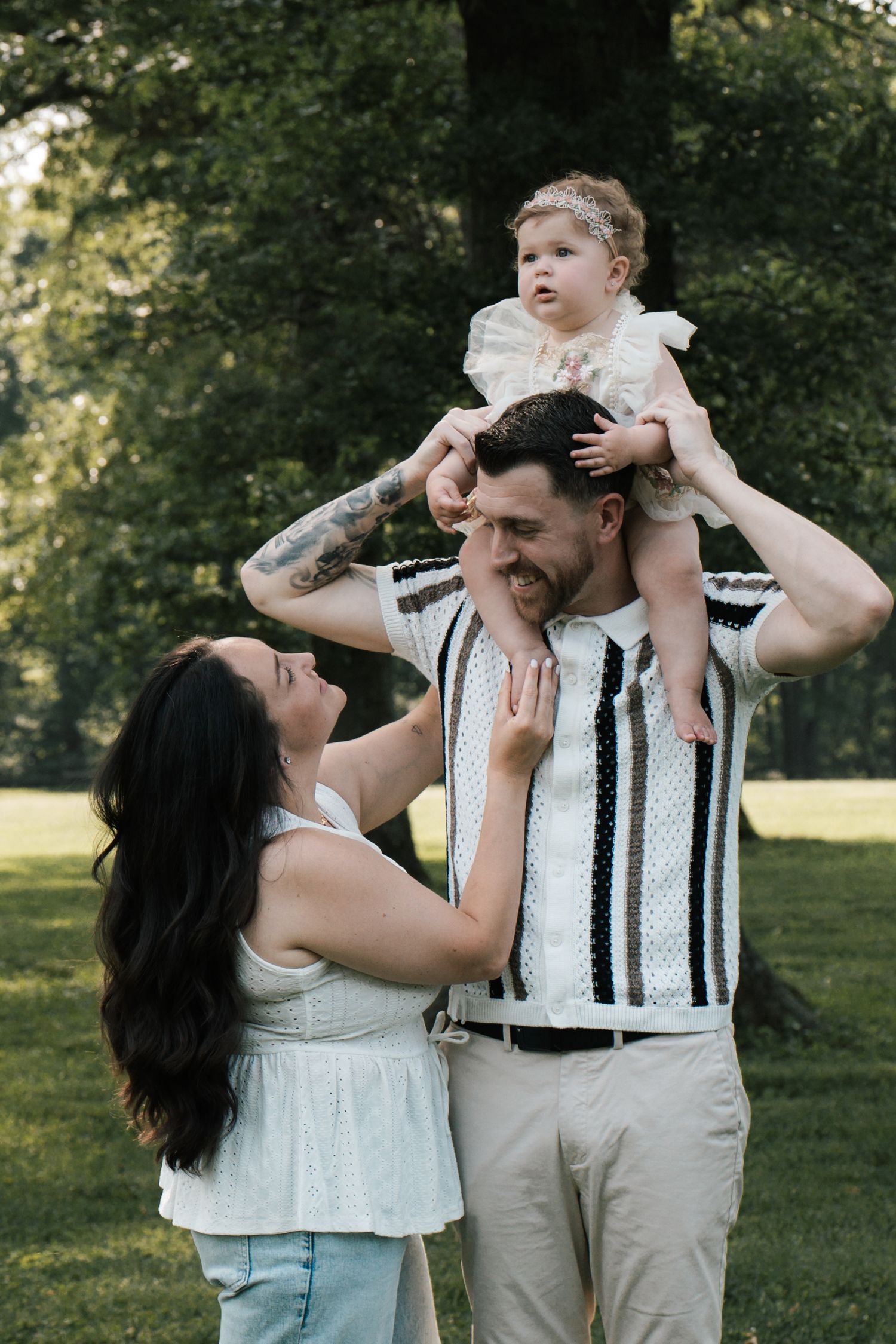 Smiling dad lifting baby girl onto shoulders while mom looks up lovingly in sunny green park.