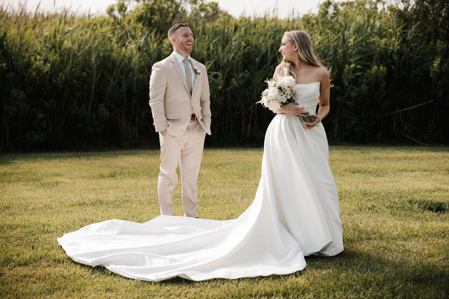 Bride in white gown with long train and groom in beige suit pose on green lawn with tall grass backdrop in New Jersey.