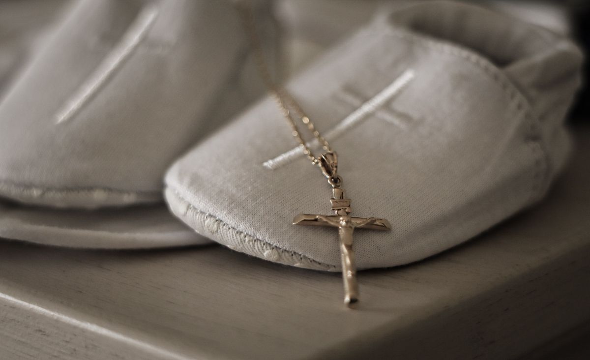 Tiny white baby shoes with a delicate gold crucifix cross necklace resting on soft fabric.