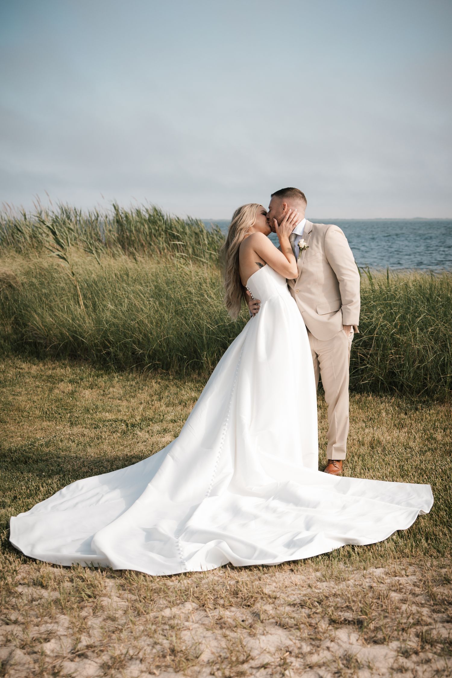 A bride and groom share a romantic kiss near coastal grass dunes with the ocean in the background in New Jersey.