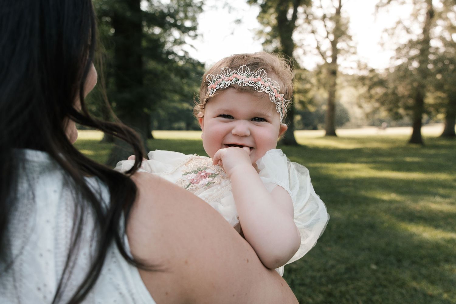 Happy chubby baby girl with floral headband smiling while being held outdoors in green park.
