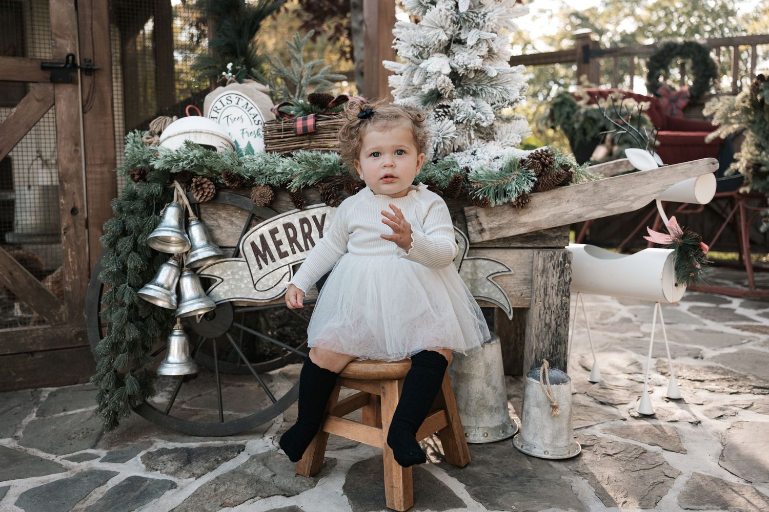 Toddler girl in white tutu dress seated on wooden stool amid festive Christmas decorations outdoors.