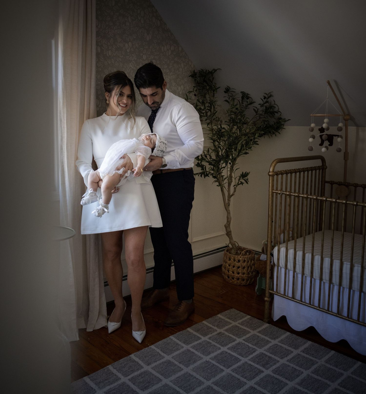 Parents in formal attire lovingly holding their baby in a white christening gown in a nursery.