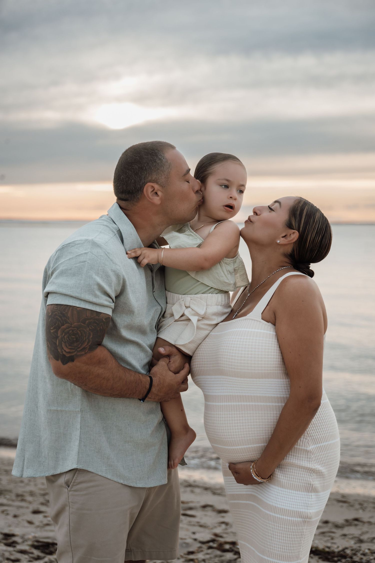 Family maternity photo of couple kissing toddler daughter on beach at sunset near ocean.