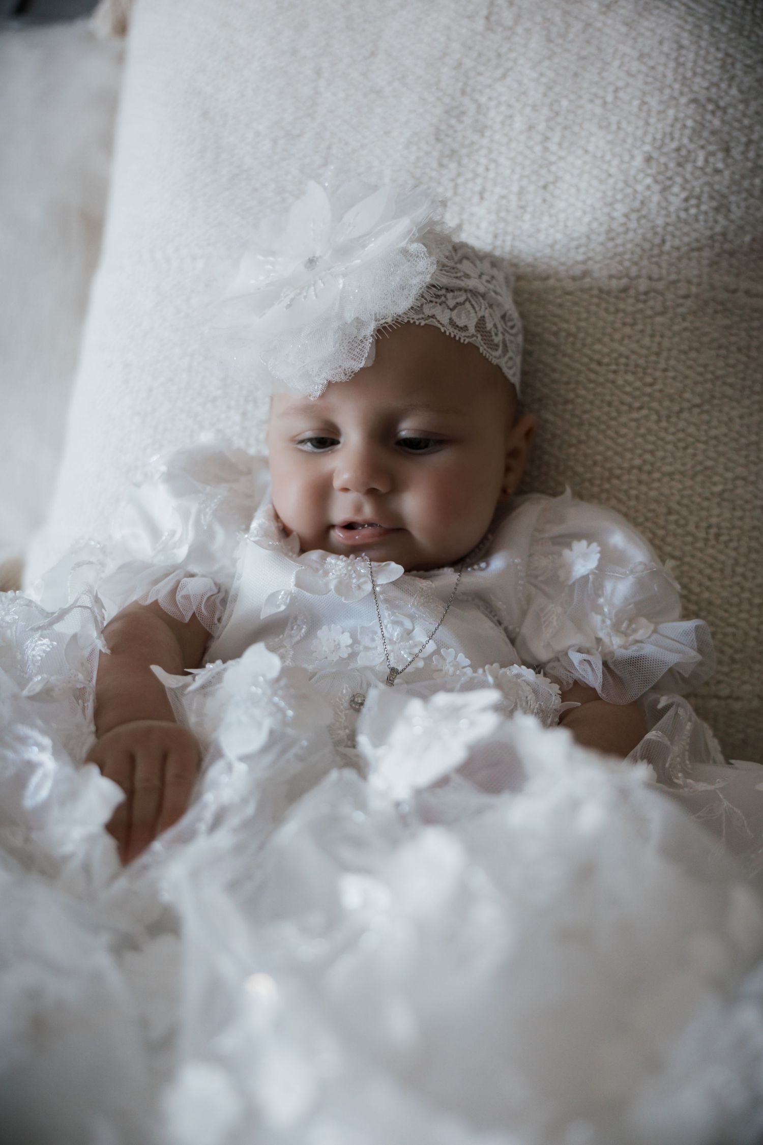 Close-up portrait of baby in white lace christening gown and floral headband resting on cream cushion.