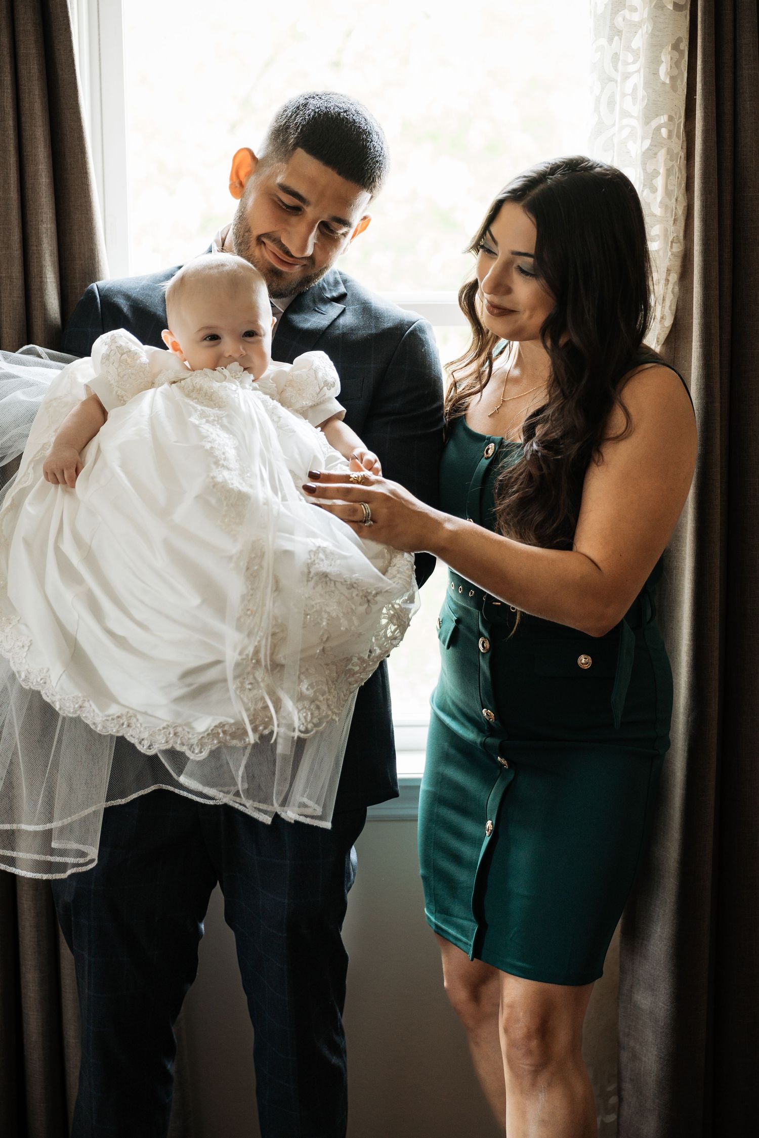 Smiling couple in formal wear holding baby dressed in white christening gown by window.