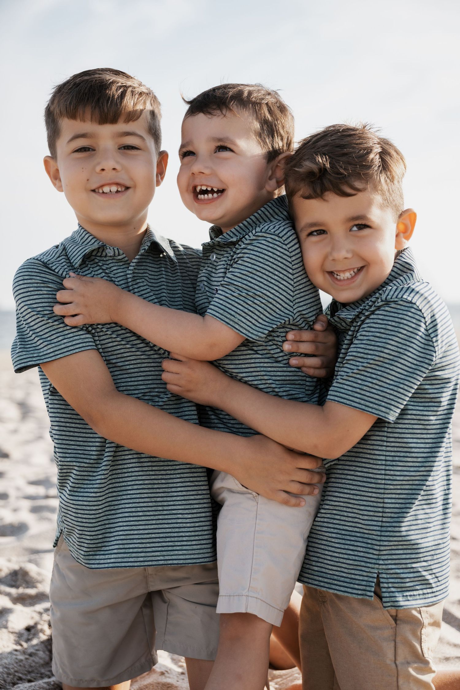 Three young brothers in matching outfits sharing a joyful group hug on a sunny beach.
