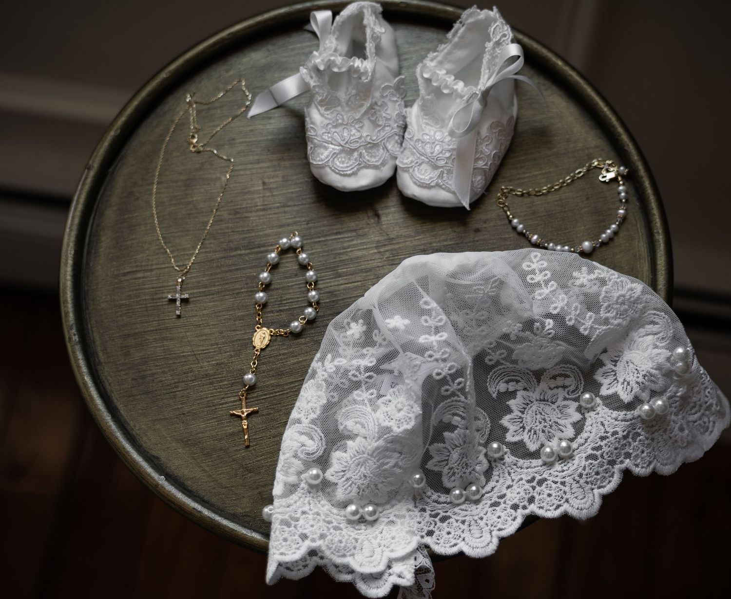 White lace baby shoes, rosary beads, gold cross necklace, and baptism accessories arranged on a vintage metal tray.
