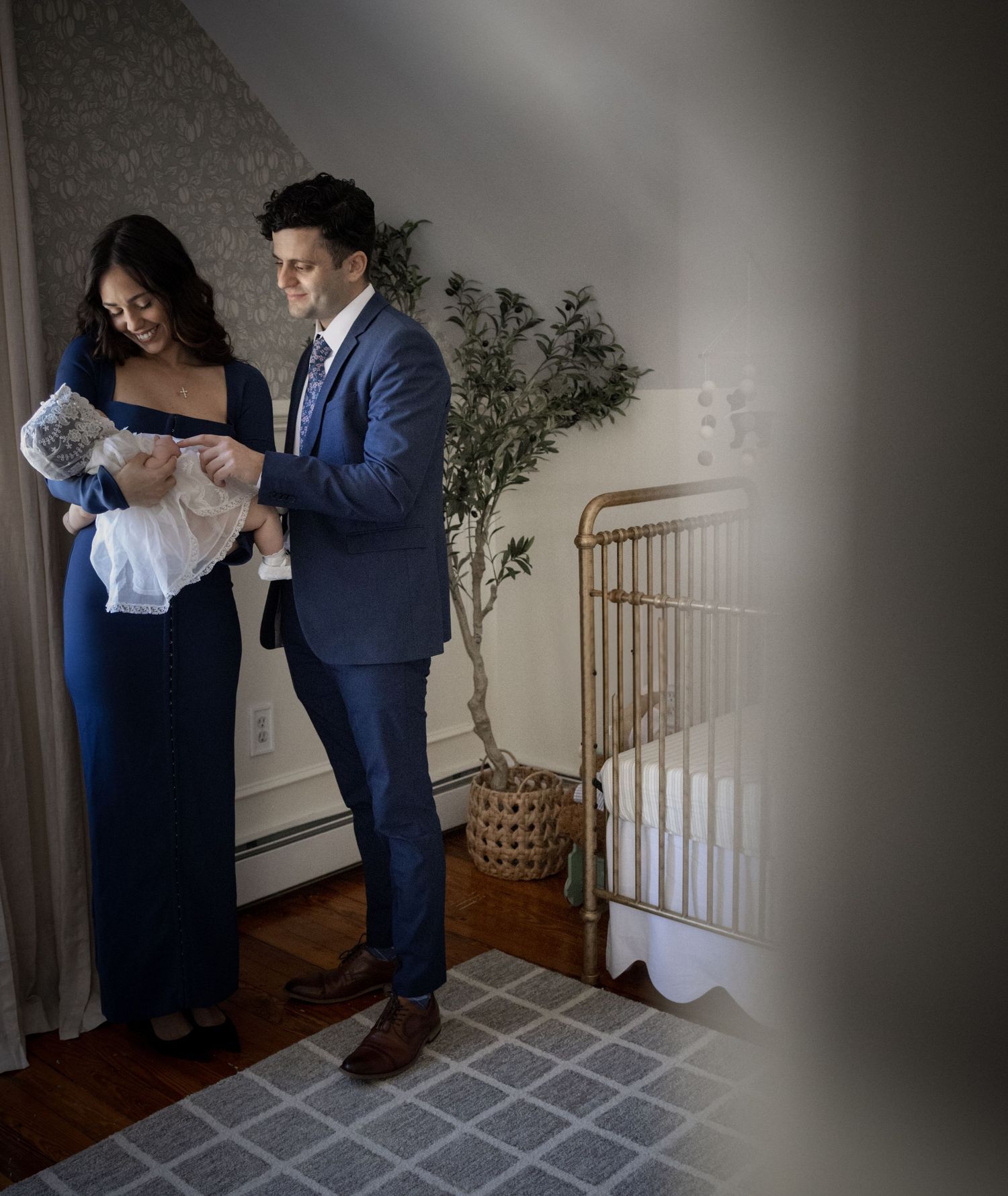 Elegant couple in formal attire preparing baby for christening ceremony beside gold crib in nursery.