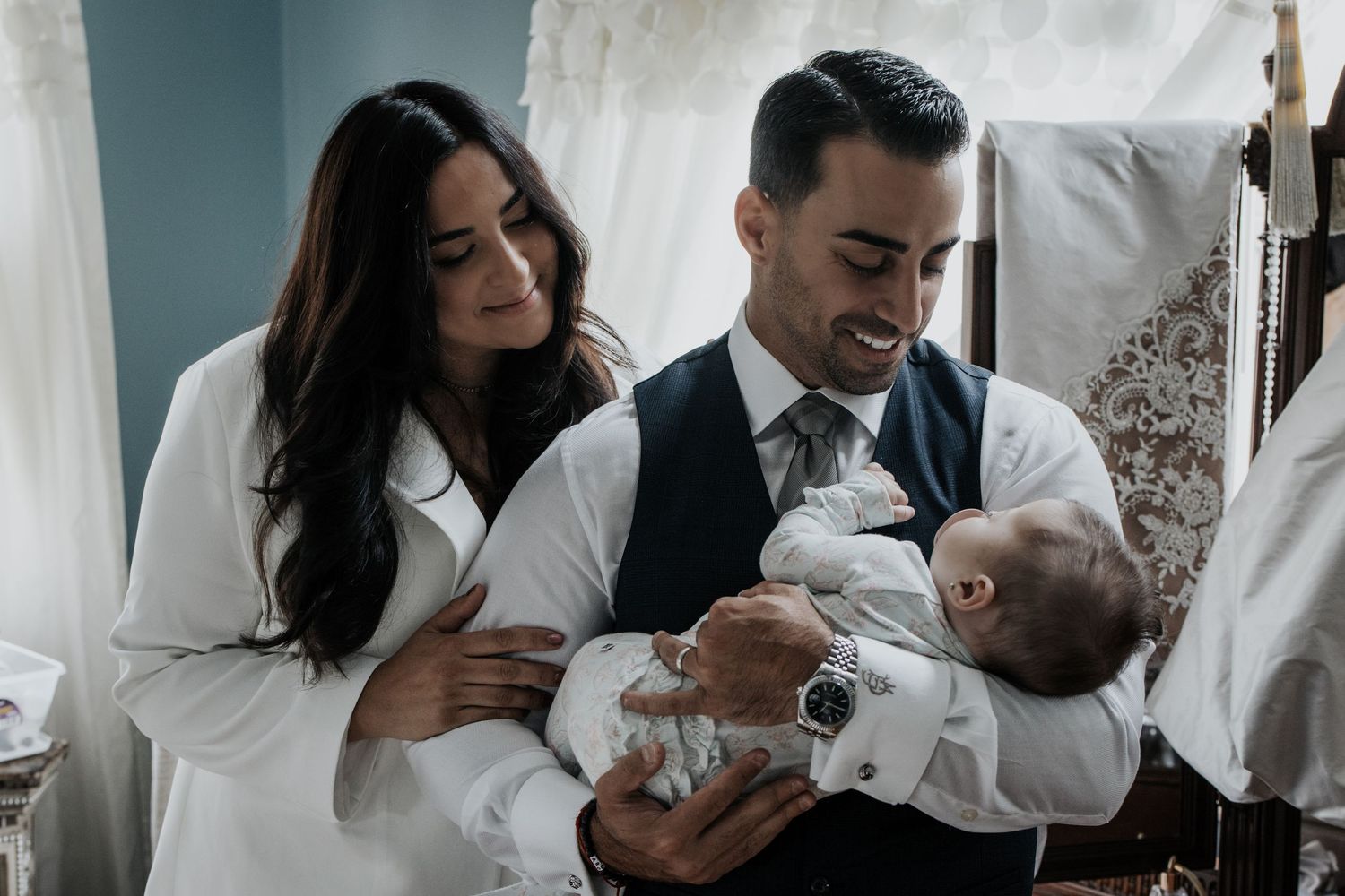 Couple in formal attire smiling at their baby during a christening ceremony indoors.
