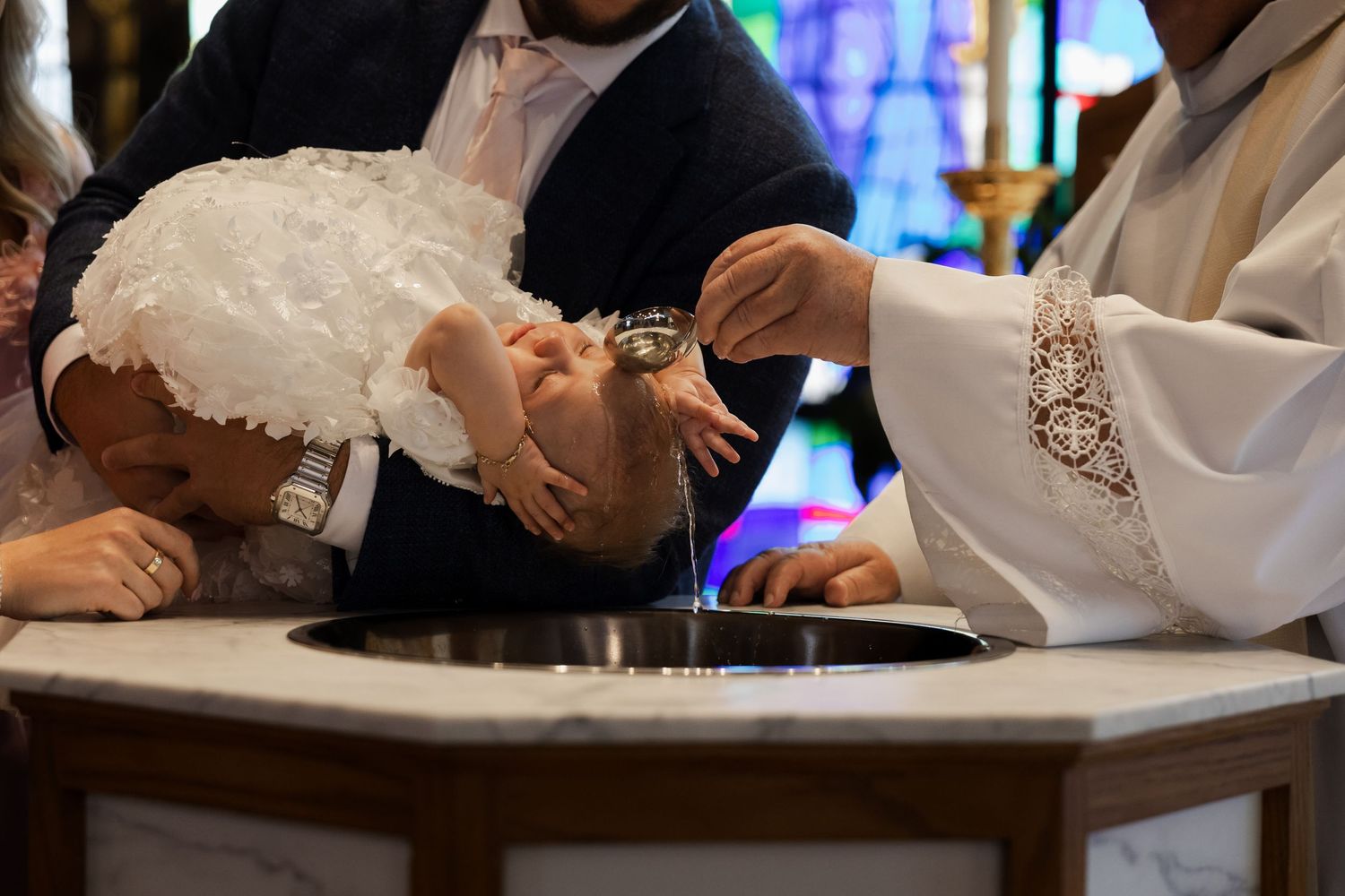 Catholic priest baptizing infant by pouring holy water over baby's head at baptismal font.