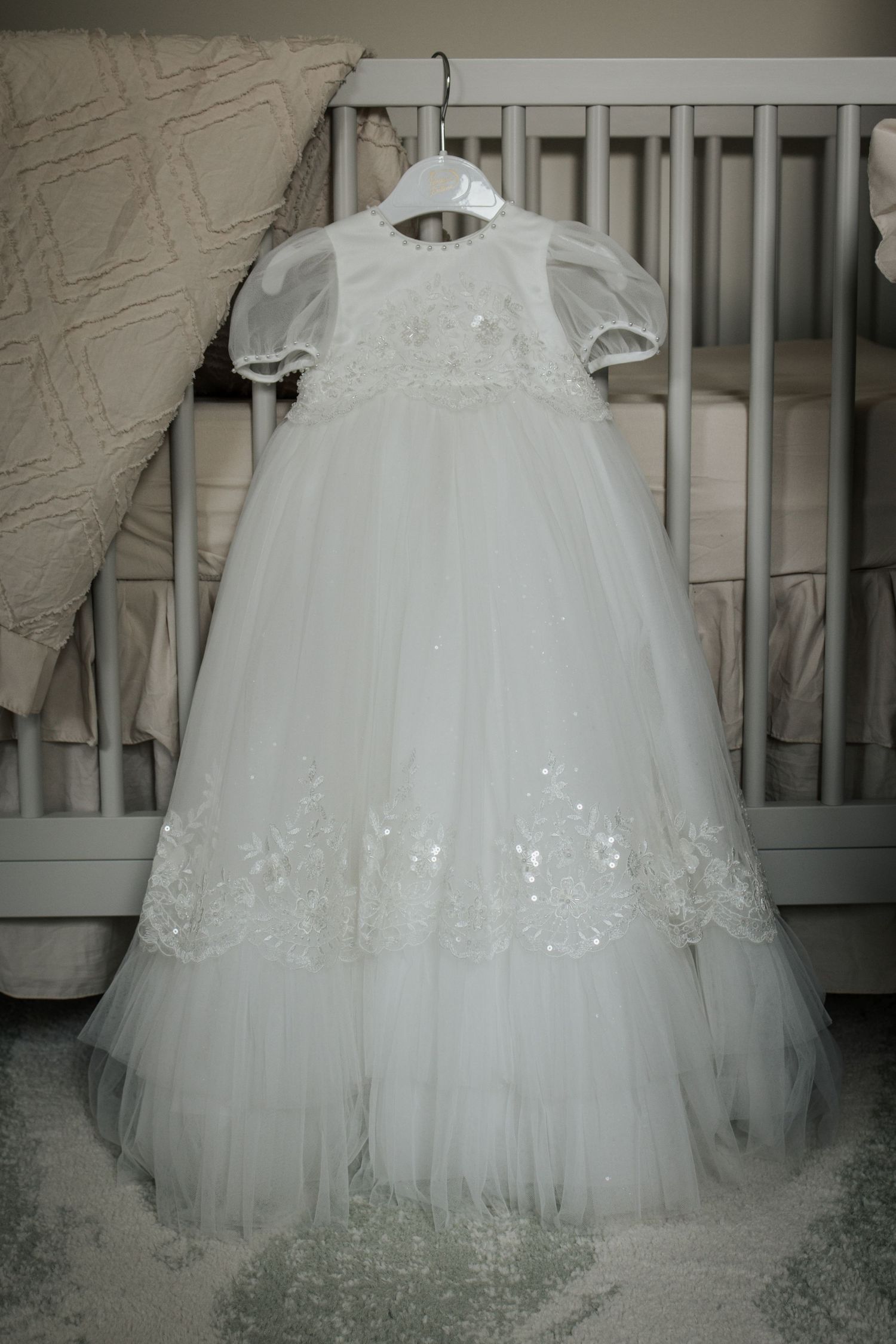 Elegant white tulle and lace baby christening gown hanging on hanger near white crib.