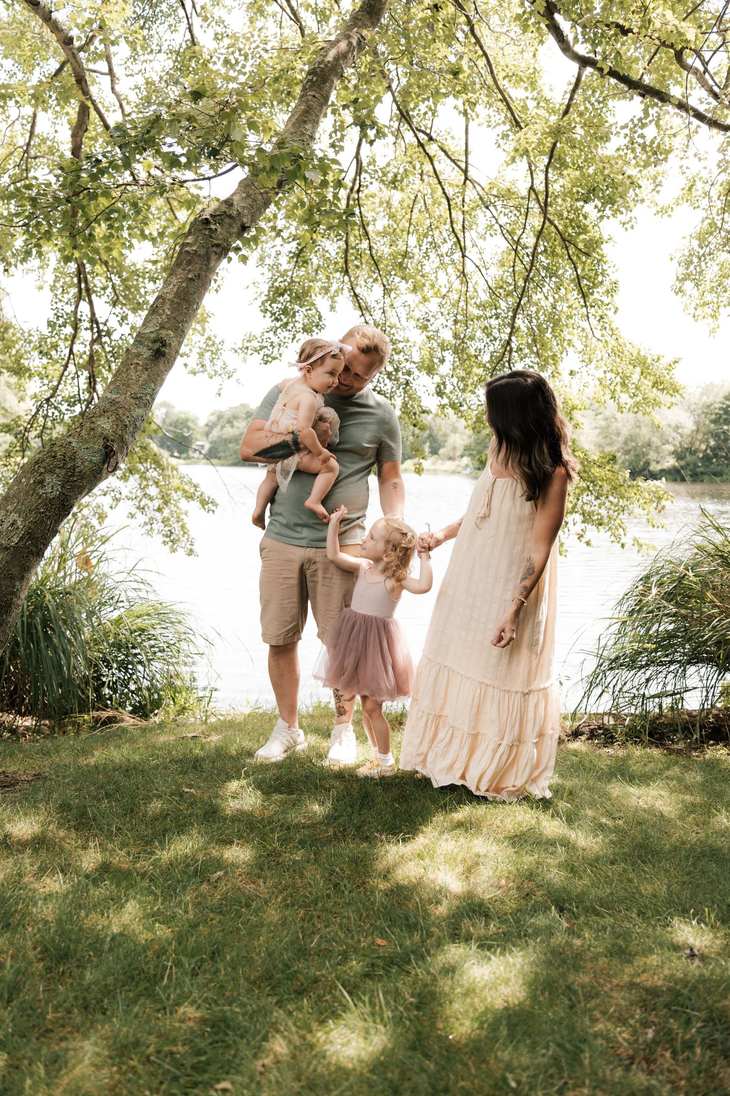 Family of four stands under tree by lake, father holding baby in pink dress.